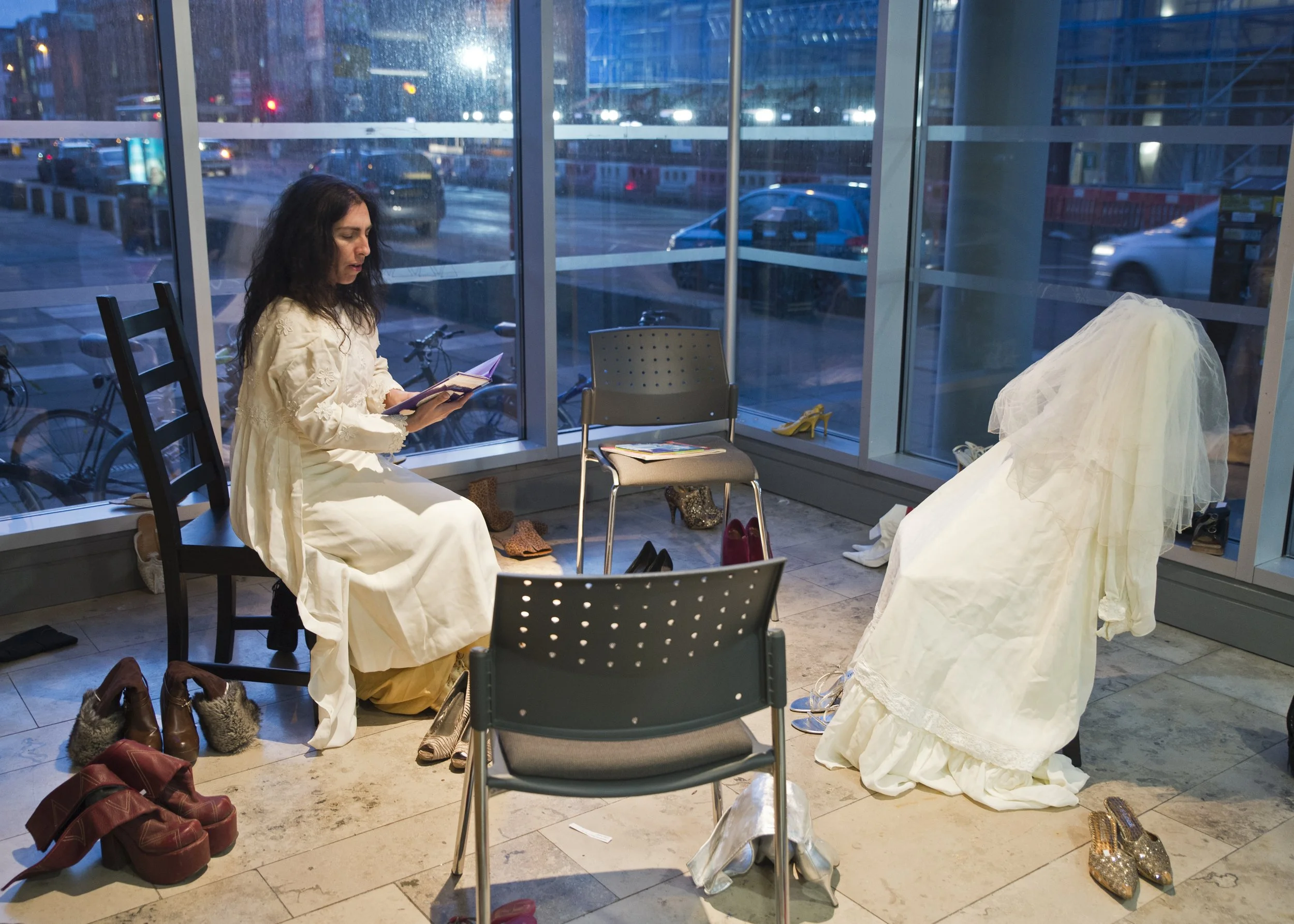 A woman sitting in a chair reading a book near a large window in an indoor space, with various shoes and bags on the floor and a dress or costume draped over a chair. The scene appears to be in a shopping mall or similar venue, with cars and building
