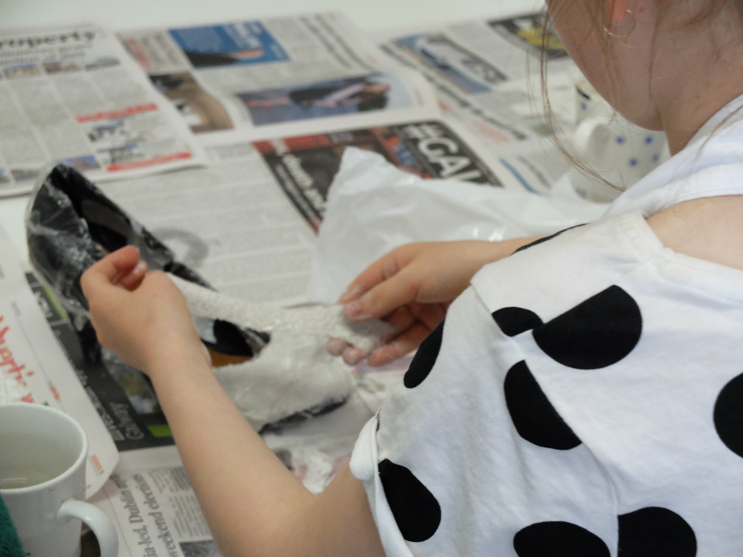 A person with light skin wearing a white shirt with black polka dots holding a black and white object, with newspapers and a white mug on the table.