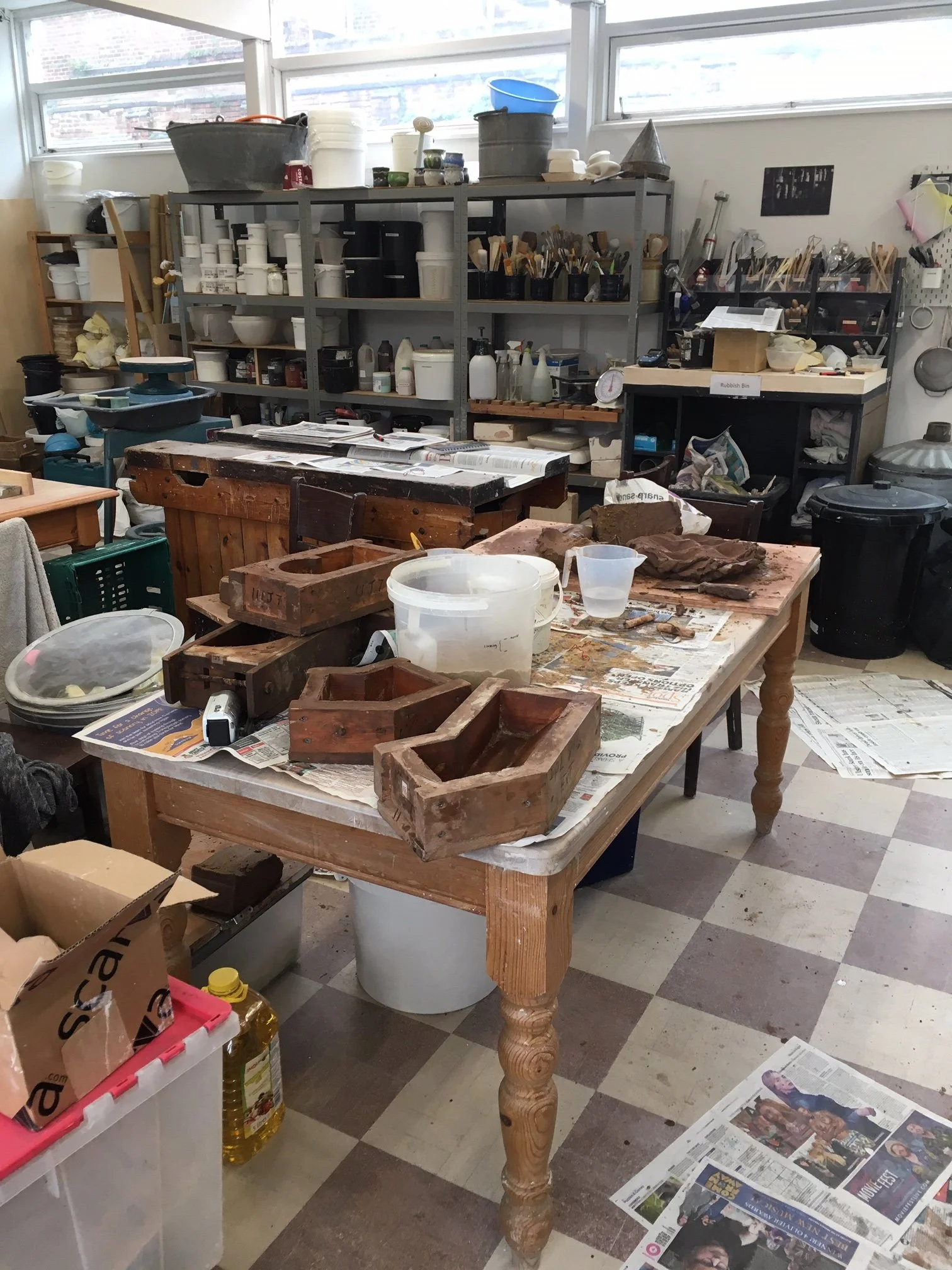 A cluttered workshop with shelves filled with art supplies, a work table with various wooden objects, newspapers on the floor, and discarded items like a plastic container, a box of oil, and various containers and tools. Large windows let in natural 