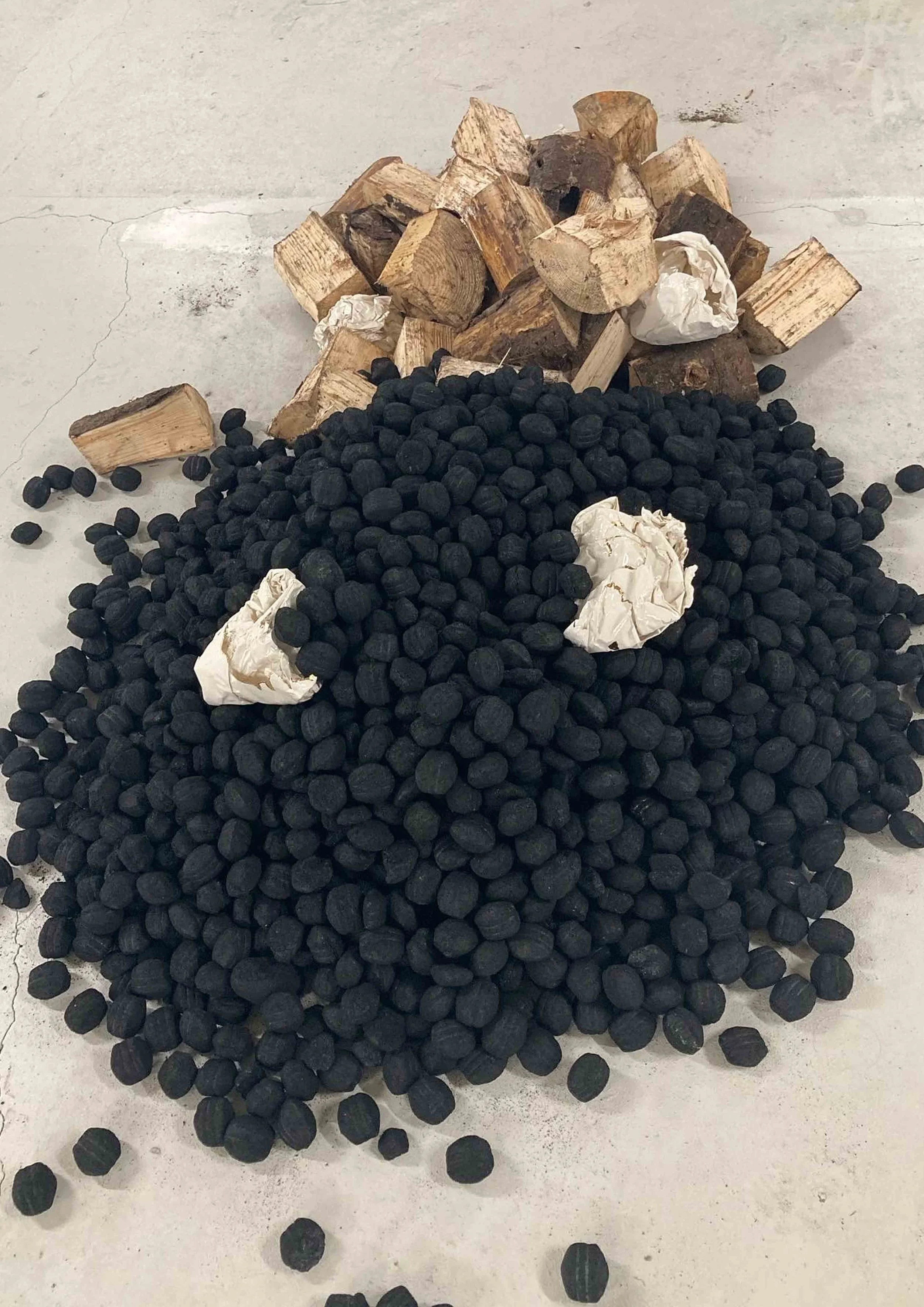 Black charcoal briquettes in a pile with some crumpled ceramic paper forms and pieces of firewood on a concrete surface.