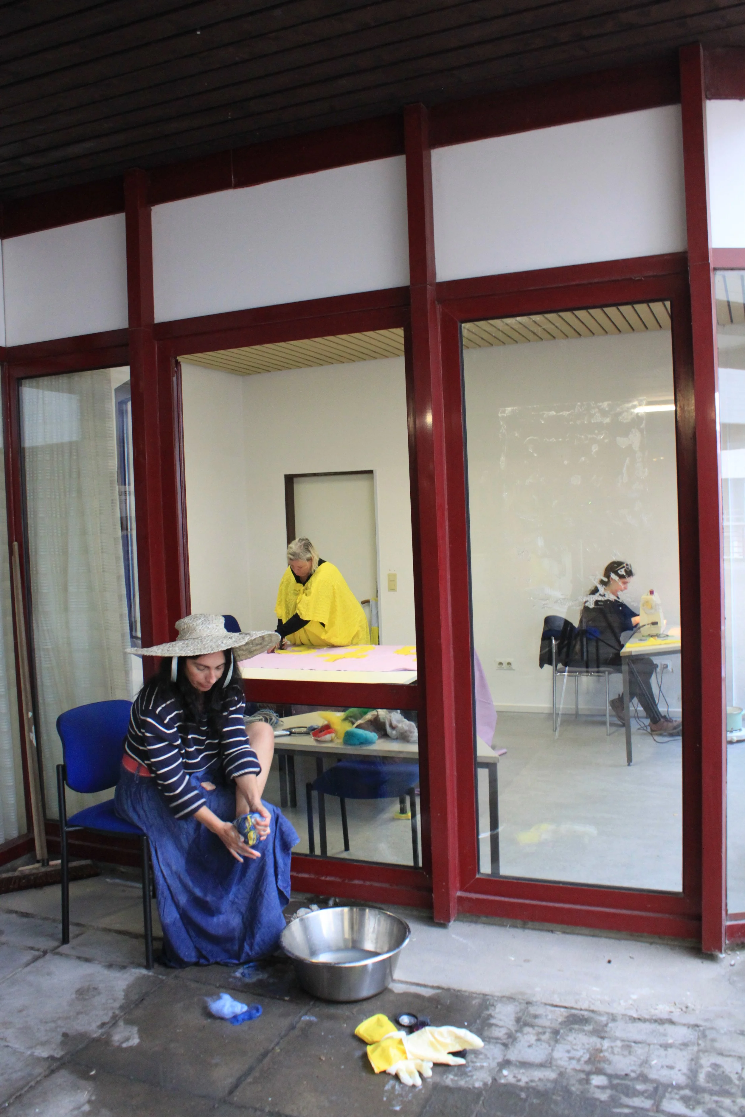 People sewing and working on craft projects inside a room, view through a glass window.