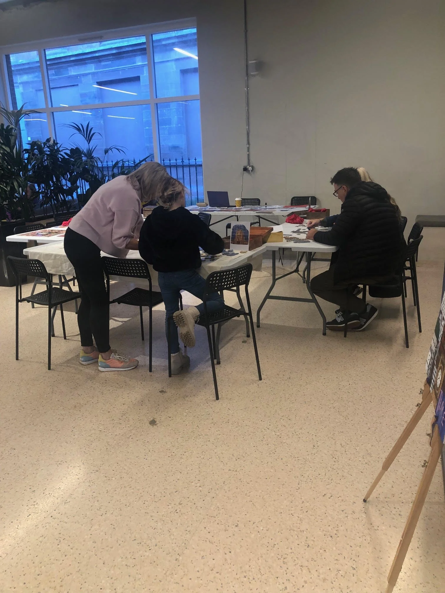 Four people are sitting at a rectangular table indoors, working with various papers and items. One person is standing, leaning over the table. The room has large windows on one side and a plain wall on the other.