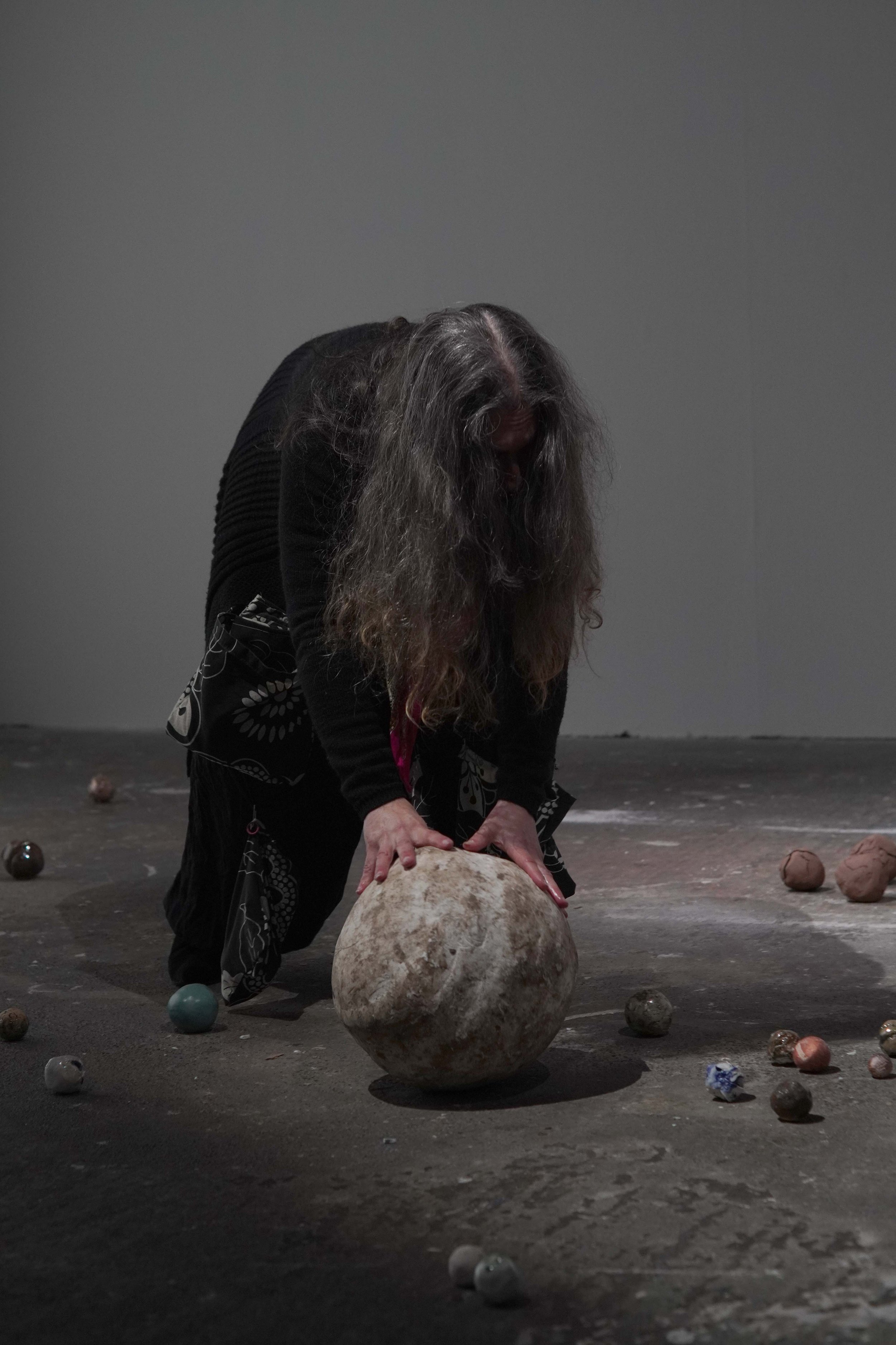 An woman with long gray hair, bowing down and touching a large stone in the center of a dimly lit room. Several smaller stones and objects are scattered on the floor around her.
