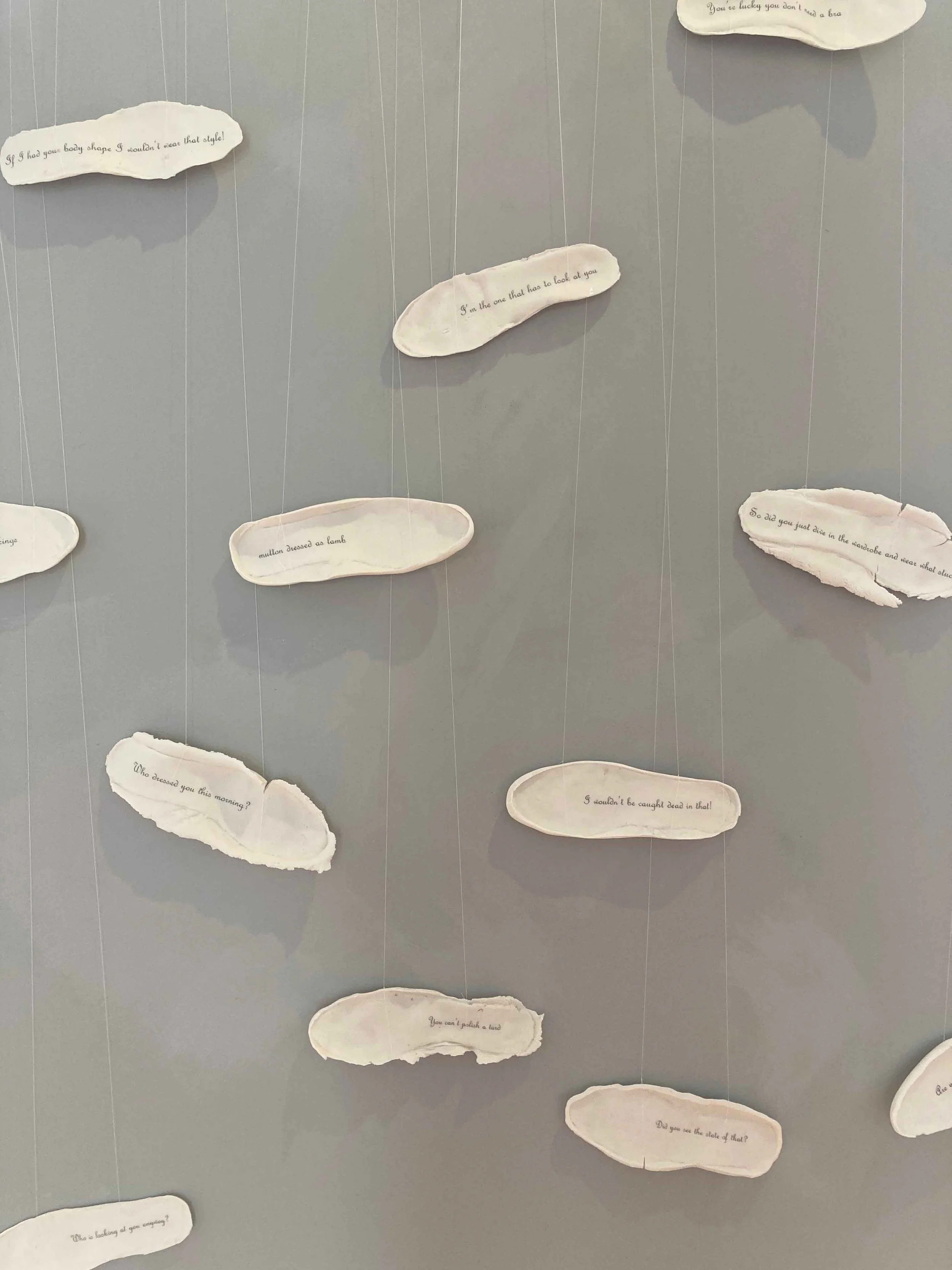 A display of ceramic footprintss with handwritten questions and statements on a light gray wall, connected by strings.