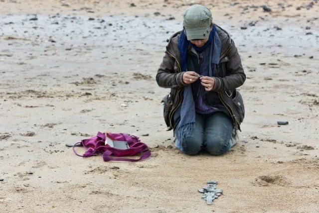 A person kneeling on a sandy beach, holding a camera or device, with a purple backpack nearby and five birds on the sand.