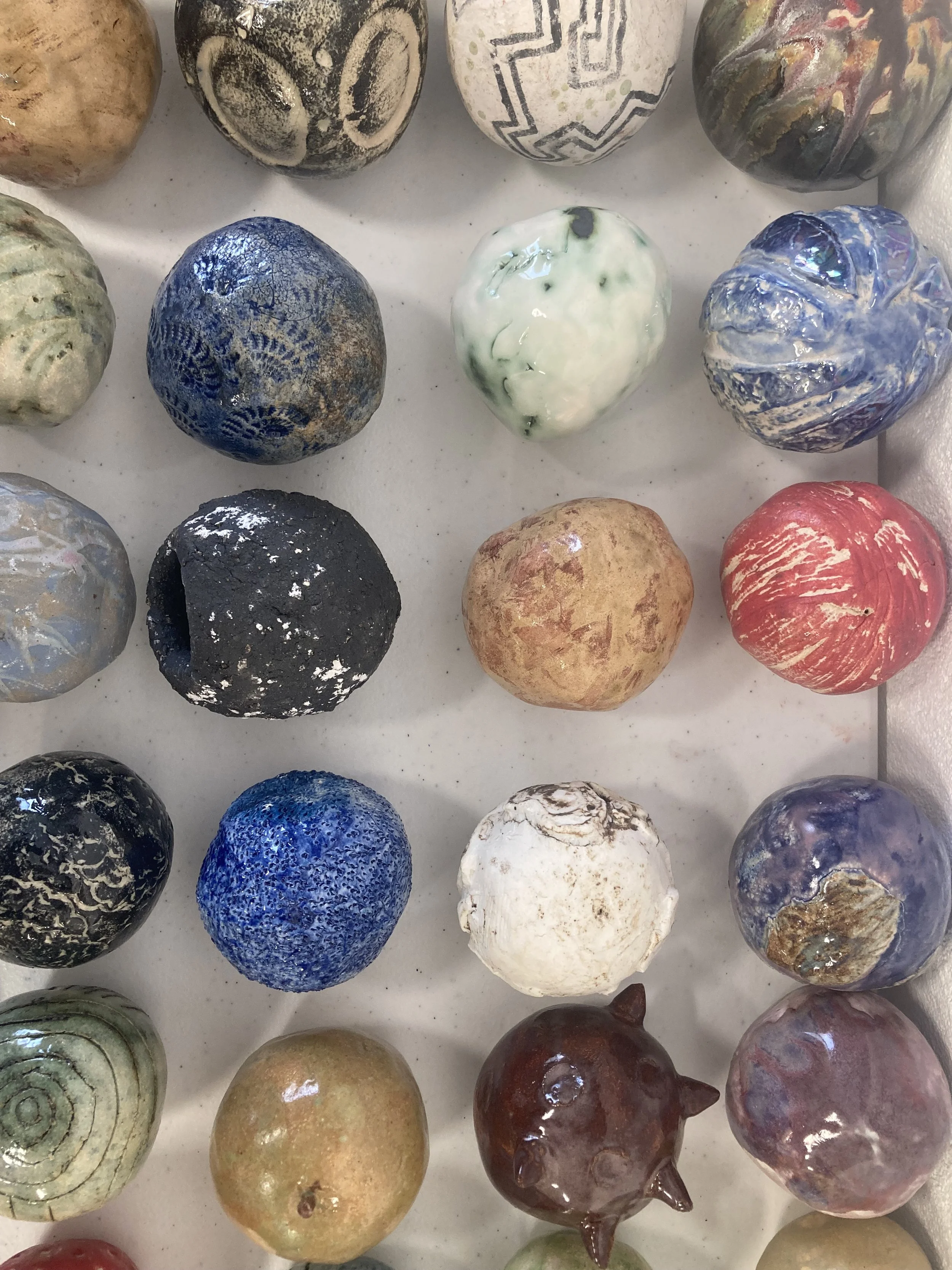 Collection of decorative painted stones with various colors and patterns, arranged in rows on a white surface.