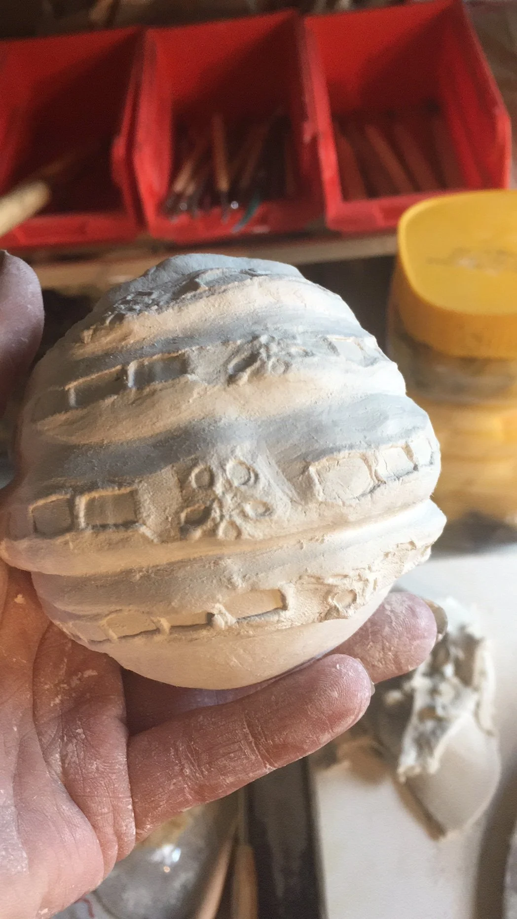 A person holding a carved stone sphere with decorative patterns, in a workspace with plastic bins and tools in the background.