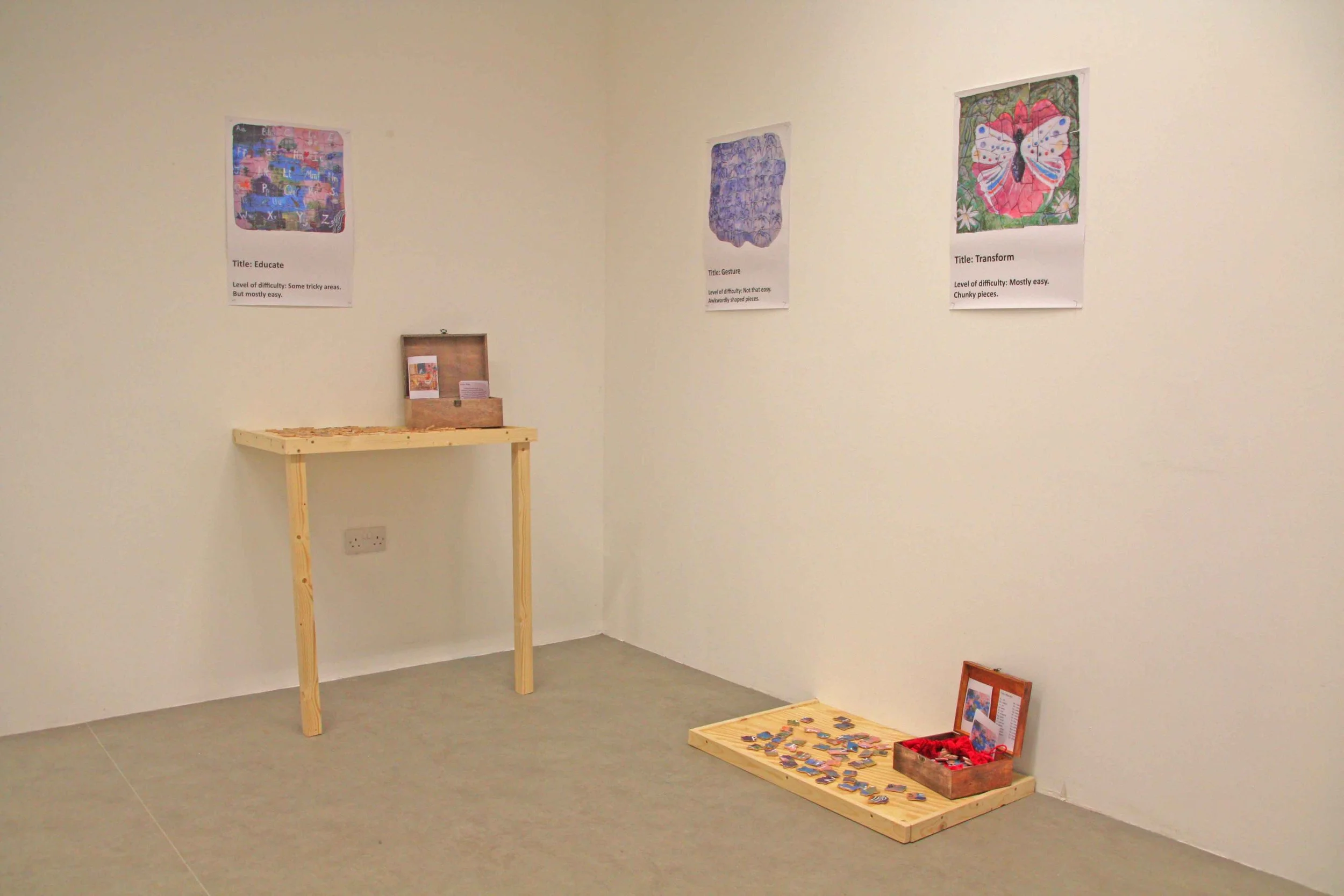 An art gallery wall with three colorful art pieces labeled 'Educate,' 'Gesture,' and 'Transform,' each with difficulty levels and descriptions. Below, there is a small wooden table with an open box of puzzles and a framed picture on the floor.