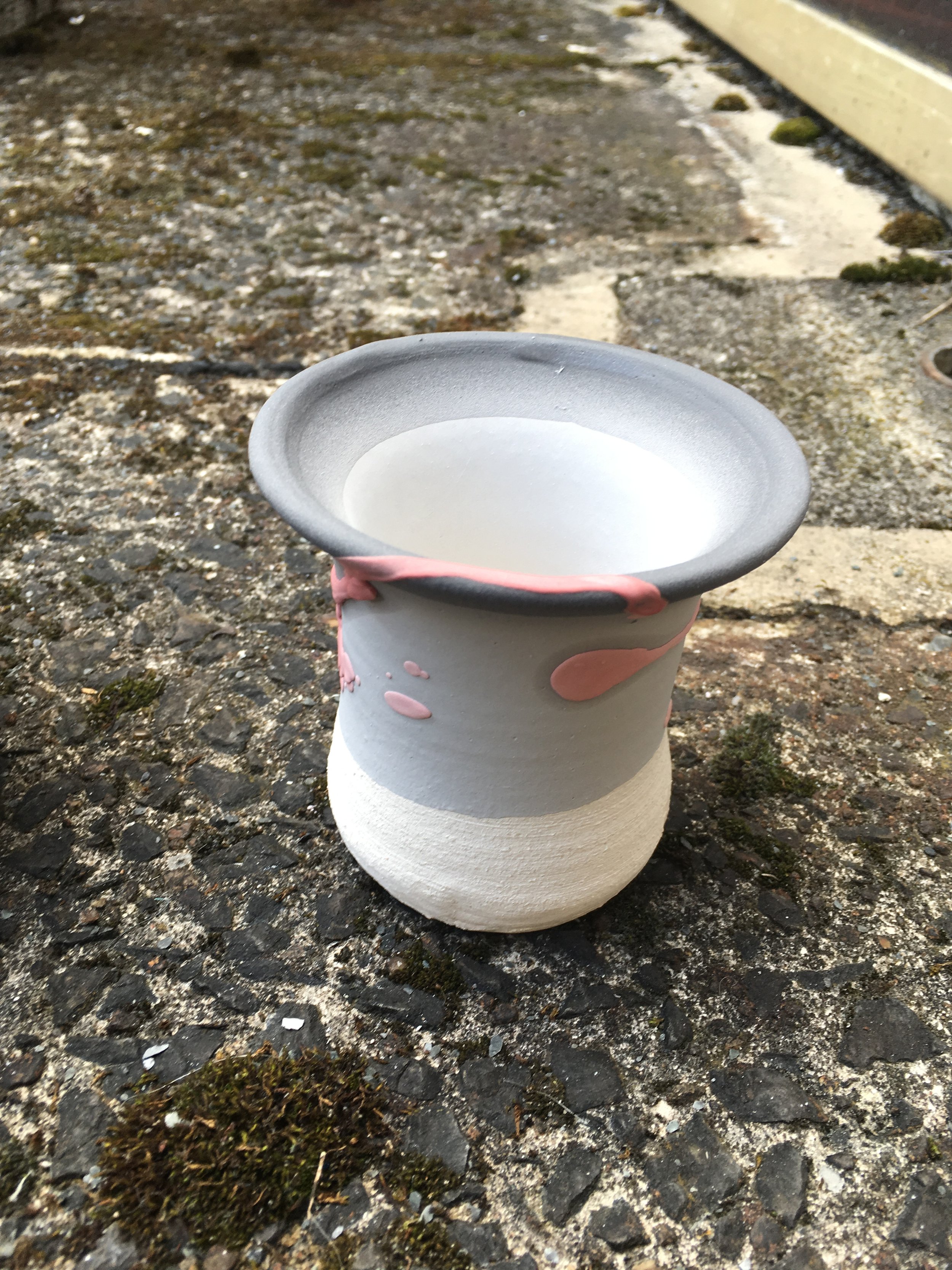 A small, round, white ceramic planter with pink accents, placed on rough concrete ground with moss and dirt, near a building's foundation.