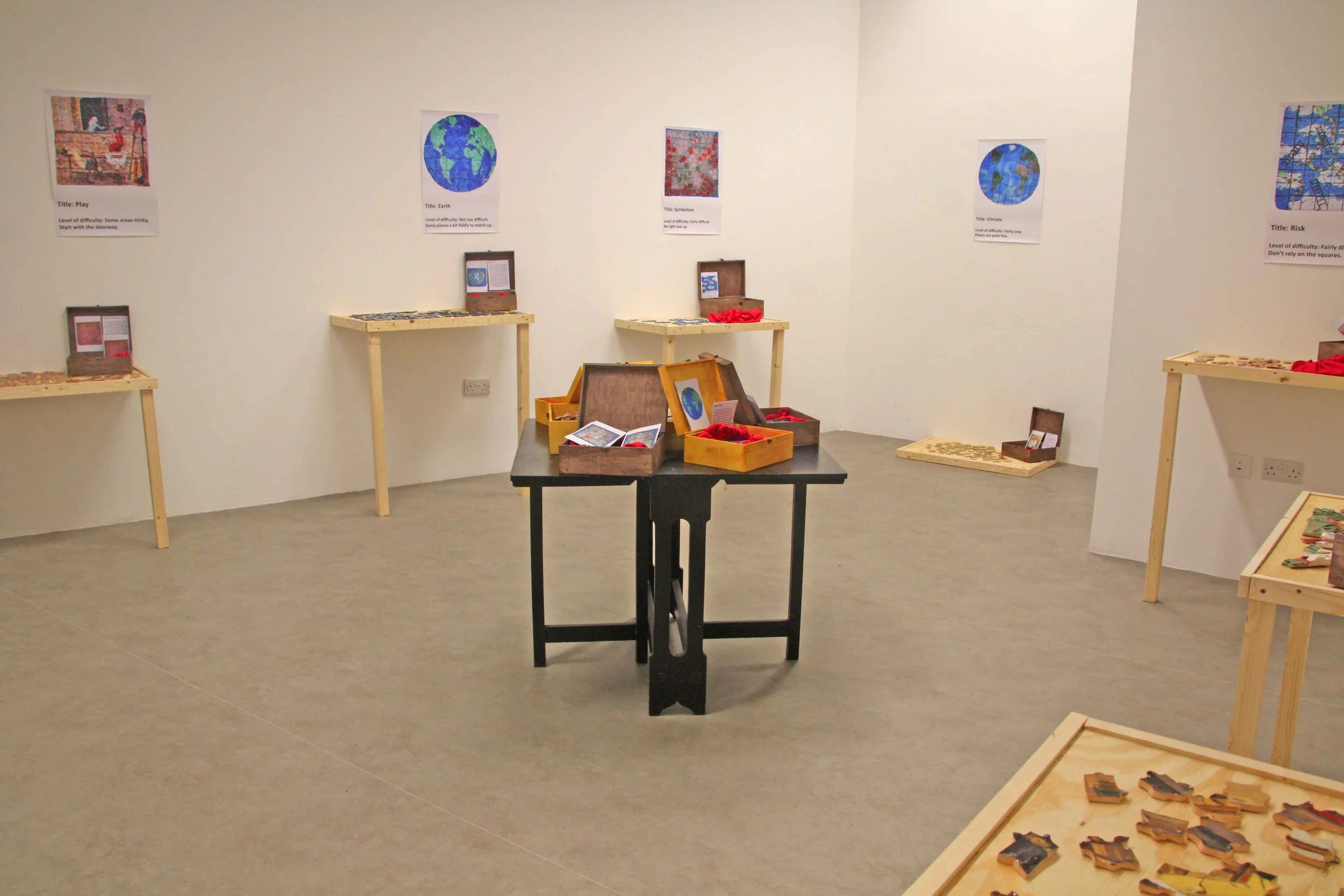 An indoor art exhibition with framed colorful images of the Earth, sky, and nature on the walls. Several wooden tables and shelves display puzzle pieces and related materials. The room has a gray floor and white walls.