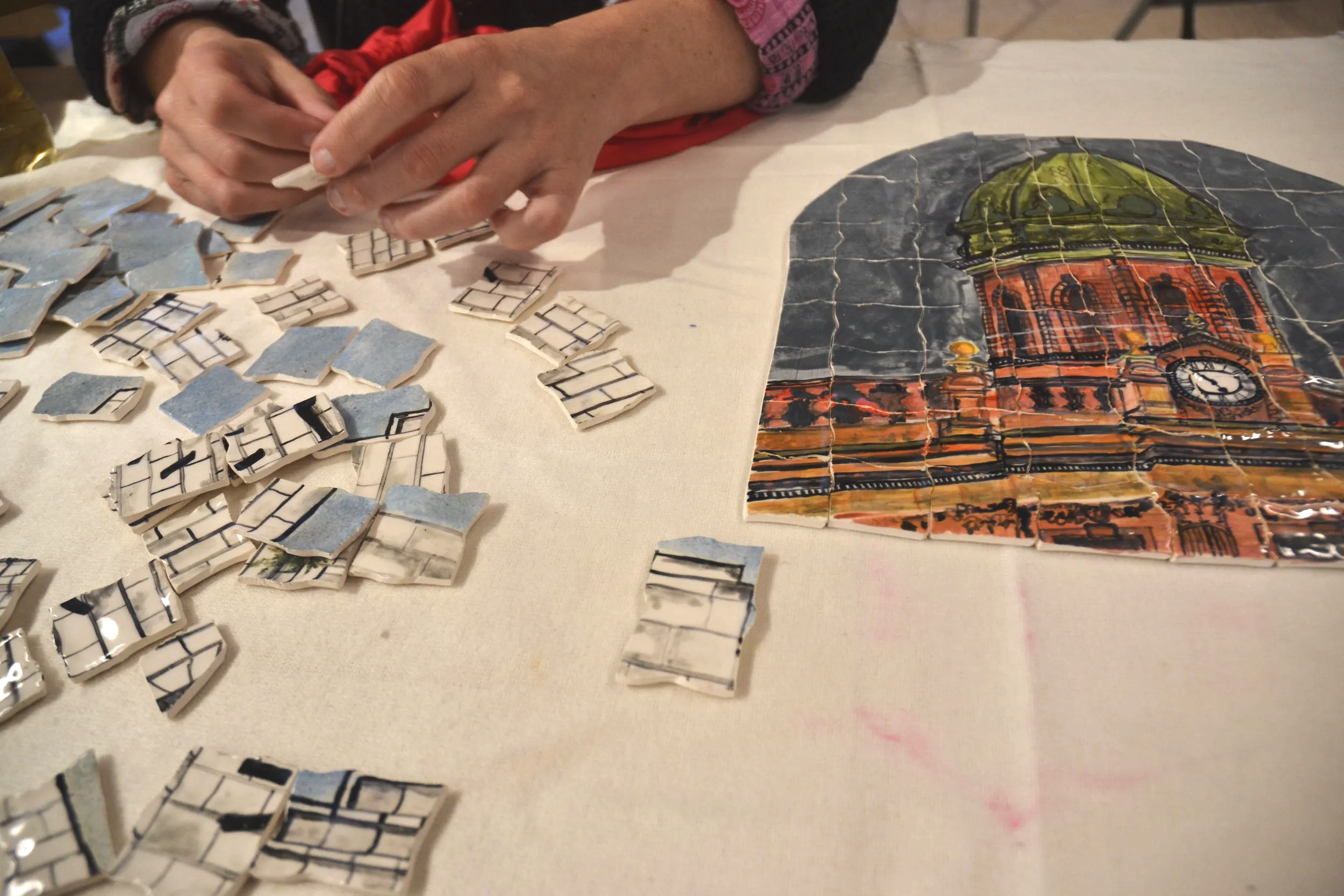 Close-up of a person working on a ceramic jigsaw puzzle depicting a clock tower, with scattered puzzle pieces on a table.