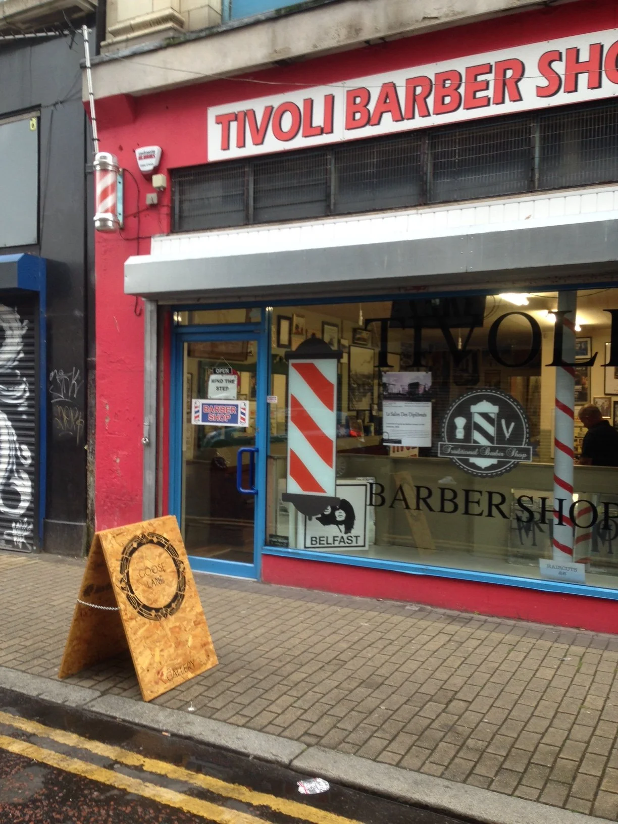 A barber shop on a city street with a red facade and large front window. The shop has a sign that reads 'Tivoli Barber Shop' in red and white. There is a barber pole outside, and a sandwich board sign on the sidewalk.