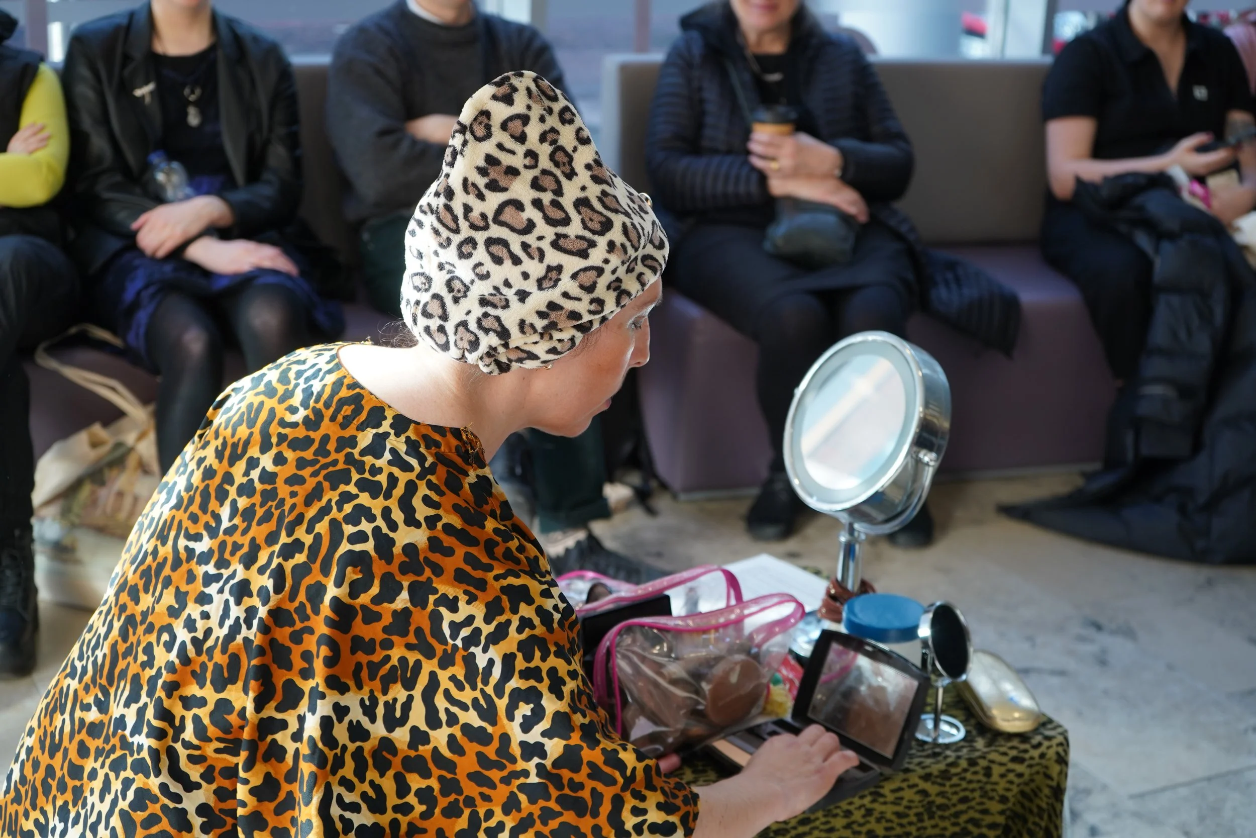 A woman wearing a leopard print headscarf and matching clothing examining a small mirror at a market or event with a group of people sitting in the background.