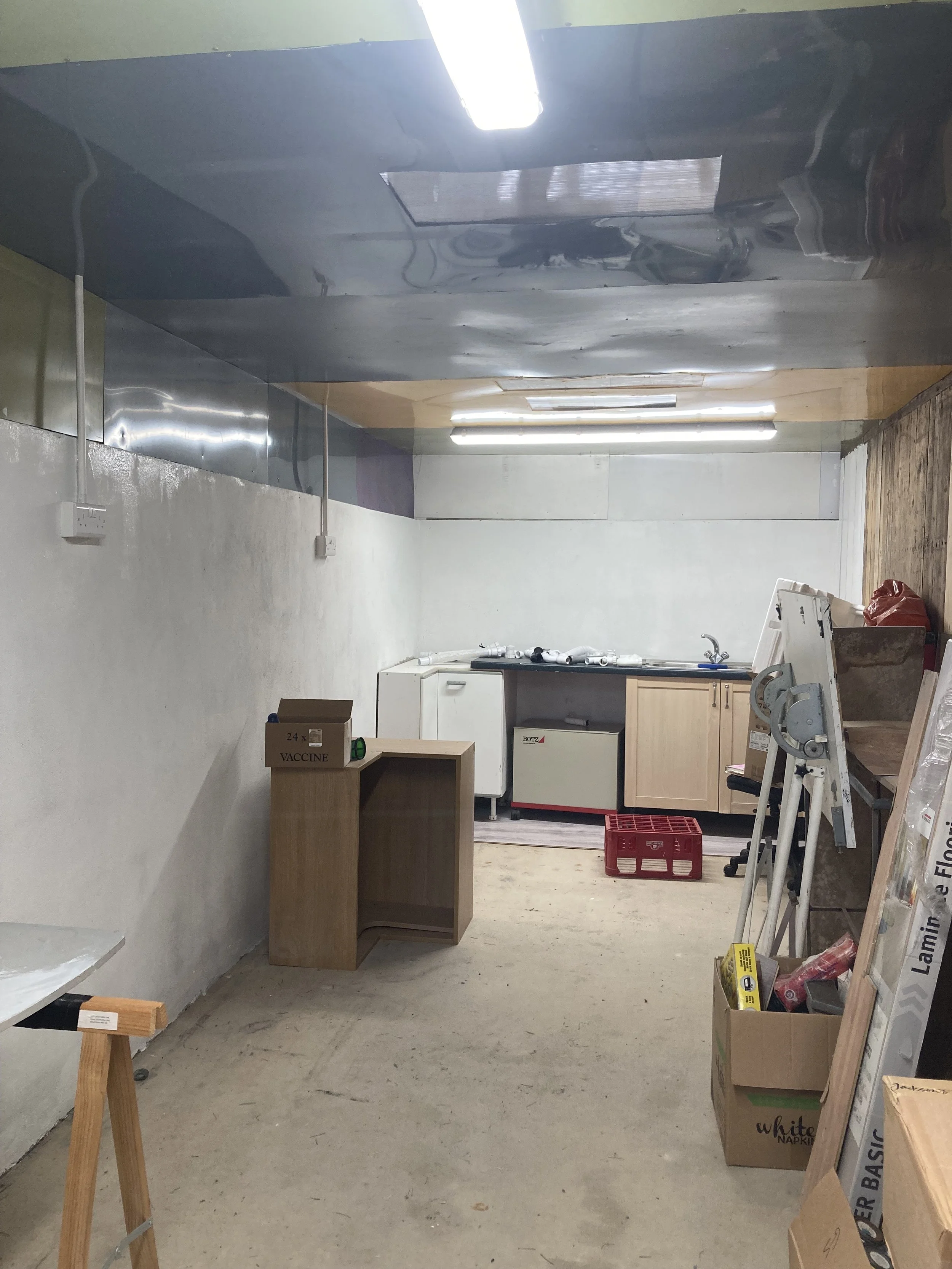 Construction room with a concrete floor, white walls, and a ceiling with fluorescent lights. There are various construction materials, a small wooden table, cardboard boxes, and a workbench with pipes and tools.