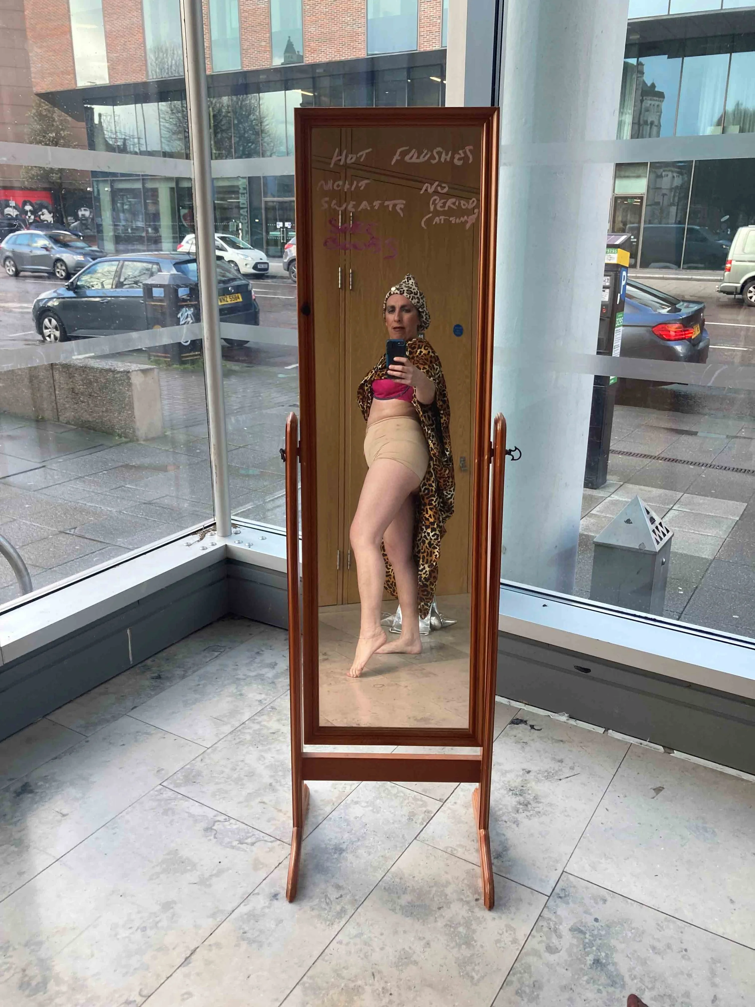A woman takes a selfie in a mirror positioned in a public space, showing her wearing a leopard print headscarf and a pink top with a beige skirt. The reflection captures her standing with one leg slightly bent and holding her phone. The mirror has no