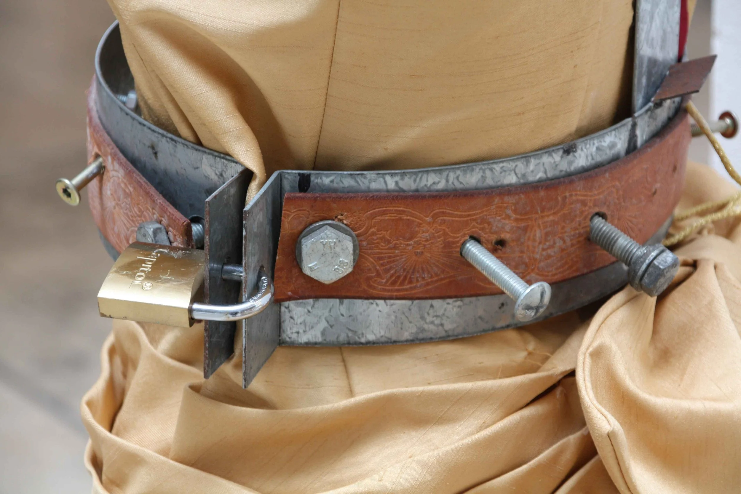 Close-up of a metallic and leather object with bolts and a lock, placed on a beige fabric surface.