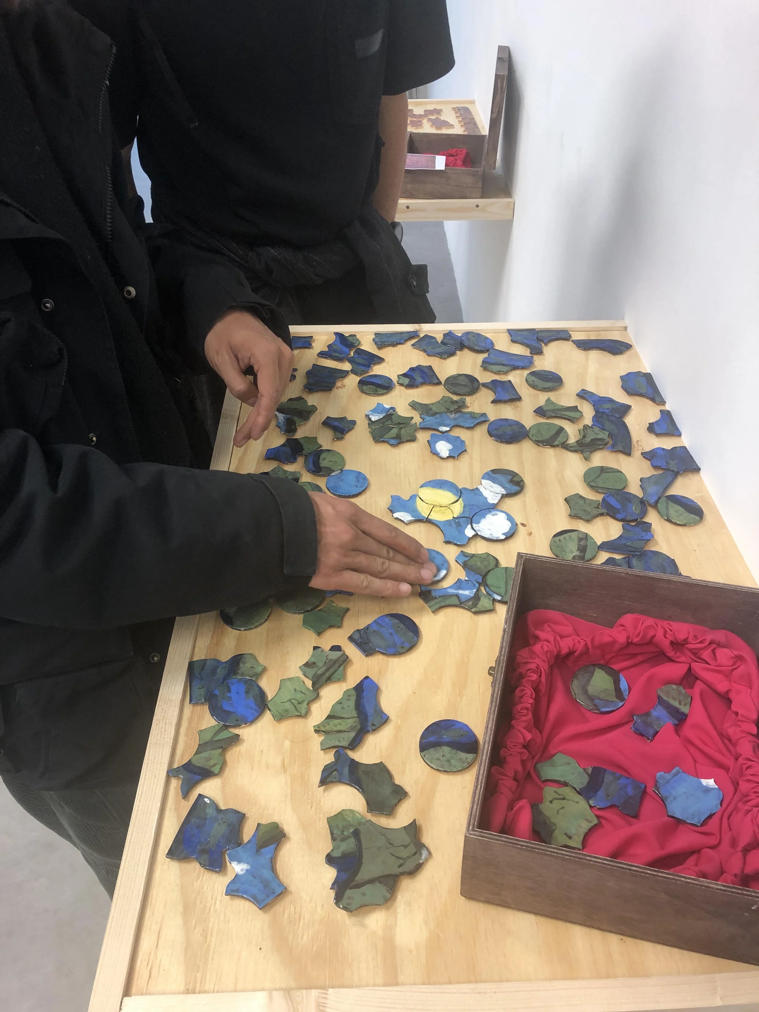 Colorful ceramic puzzle pieces arranged on a wooden table, with a person's hand placing one piece, and a red fabric-lined box filled with more puzzle pieces next to it.