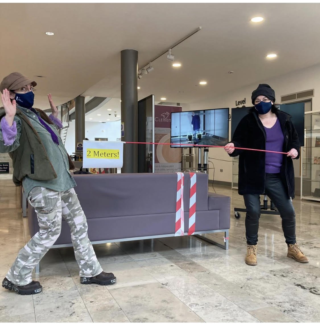 Two women wearing masks and jackets are maintaining a 2-meter distance, holding a pink rope in an indoor public space. A sign reads '2 Meters!' and a TV screen displays a blue-toned image of a person.