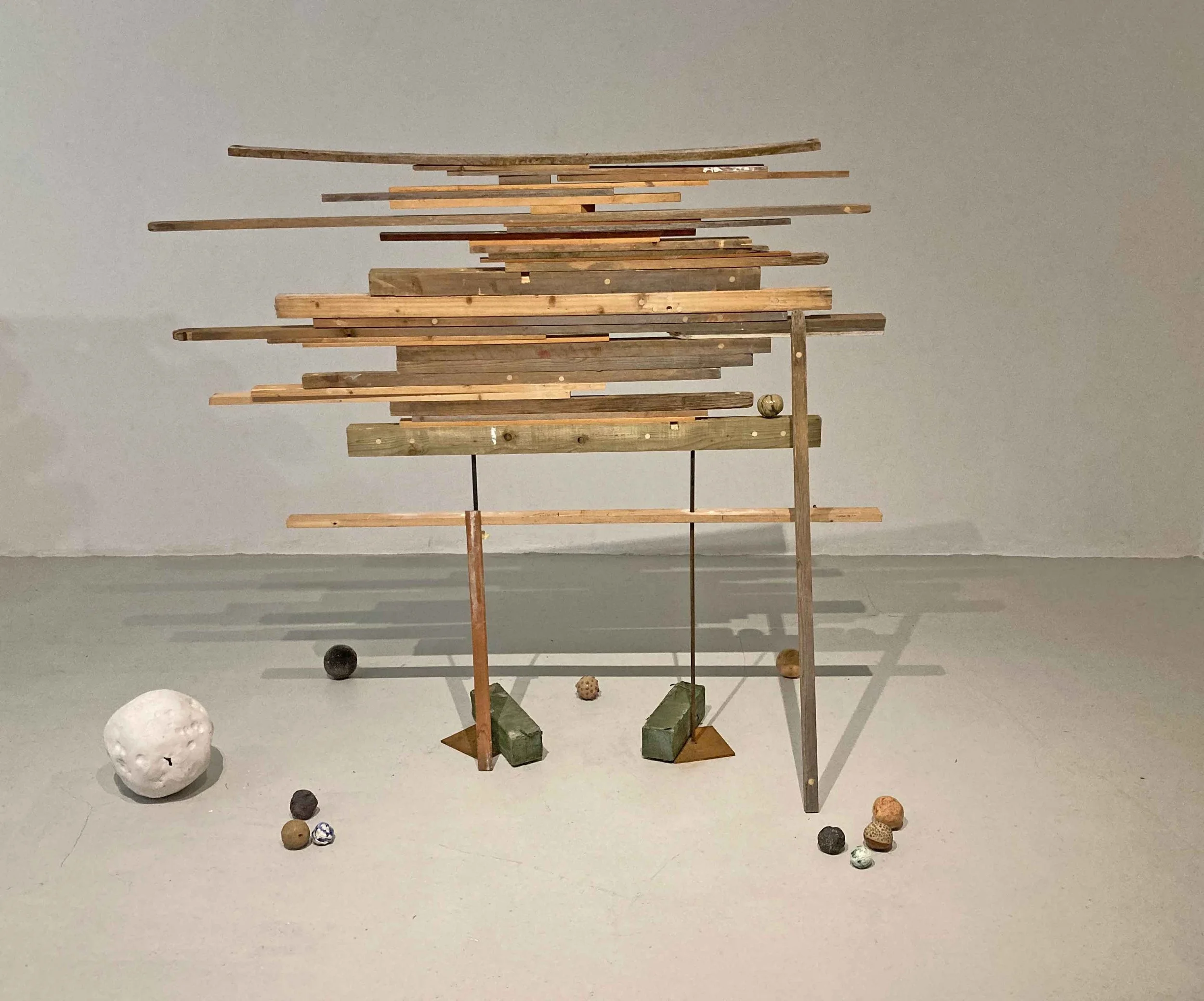 An abstract sculpture made of wooden planks arranged in a layered, horizontal pattern on a metal frame, with various small decorative objects and balls placed around it, against a plain light gray wall.