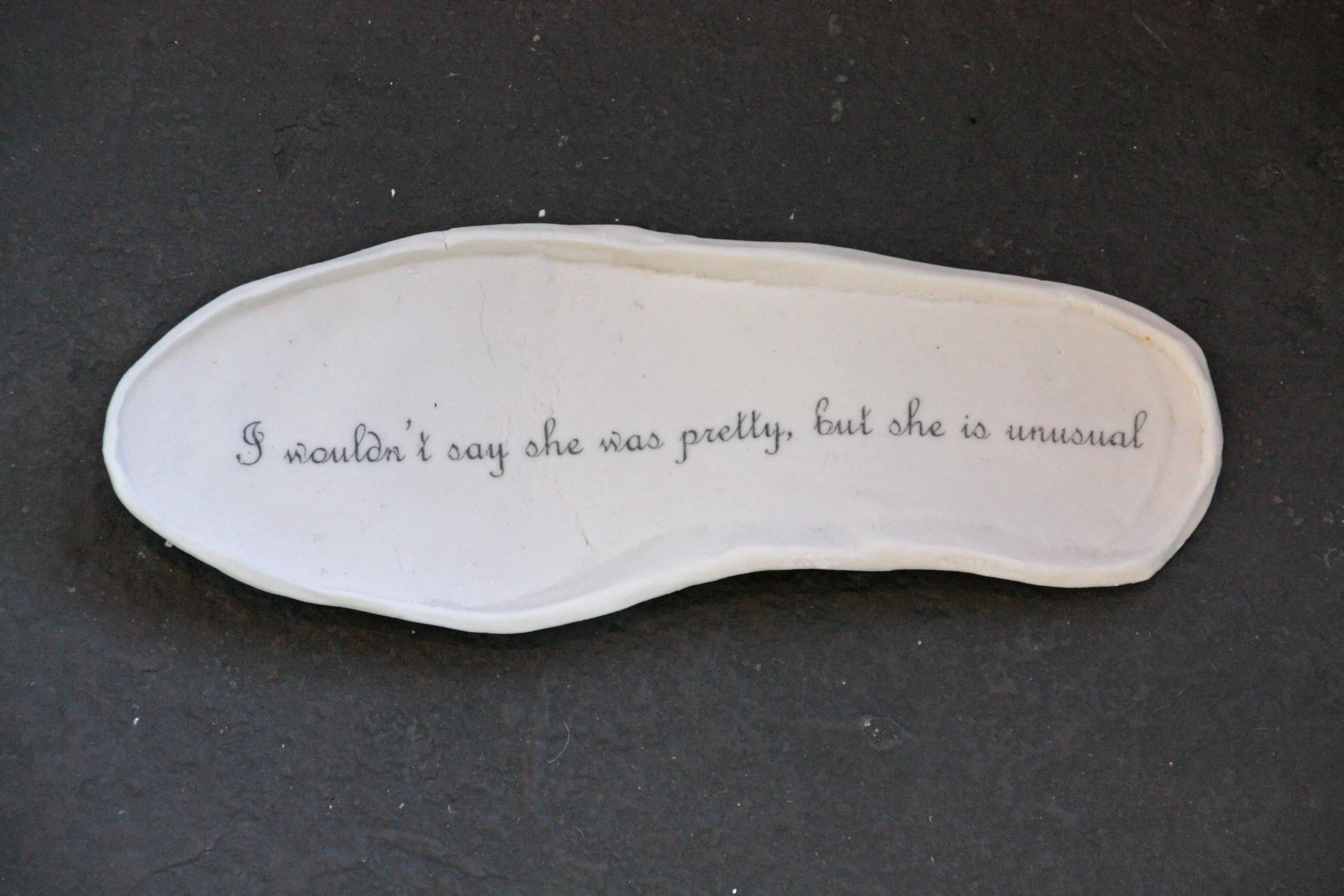 A white ceramic footprintwith black handwritten text that reads, "I wouldn't say she was pretty, but she is unusual," placed on a black surface.