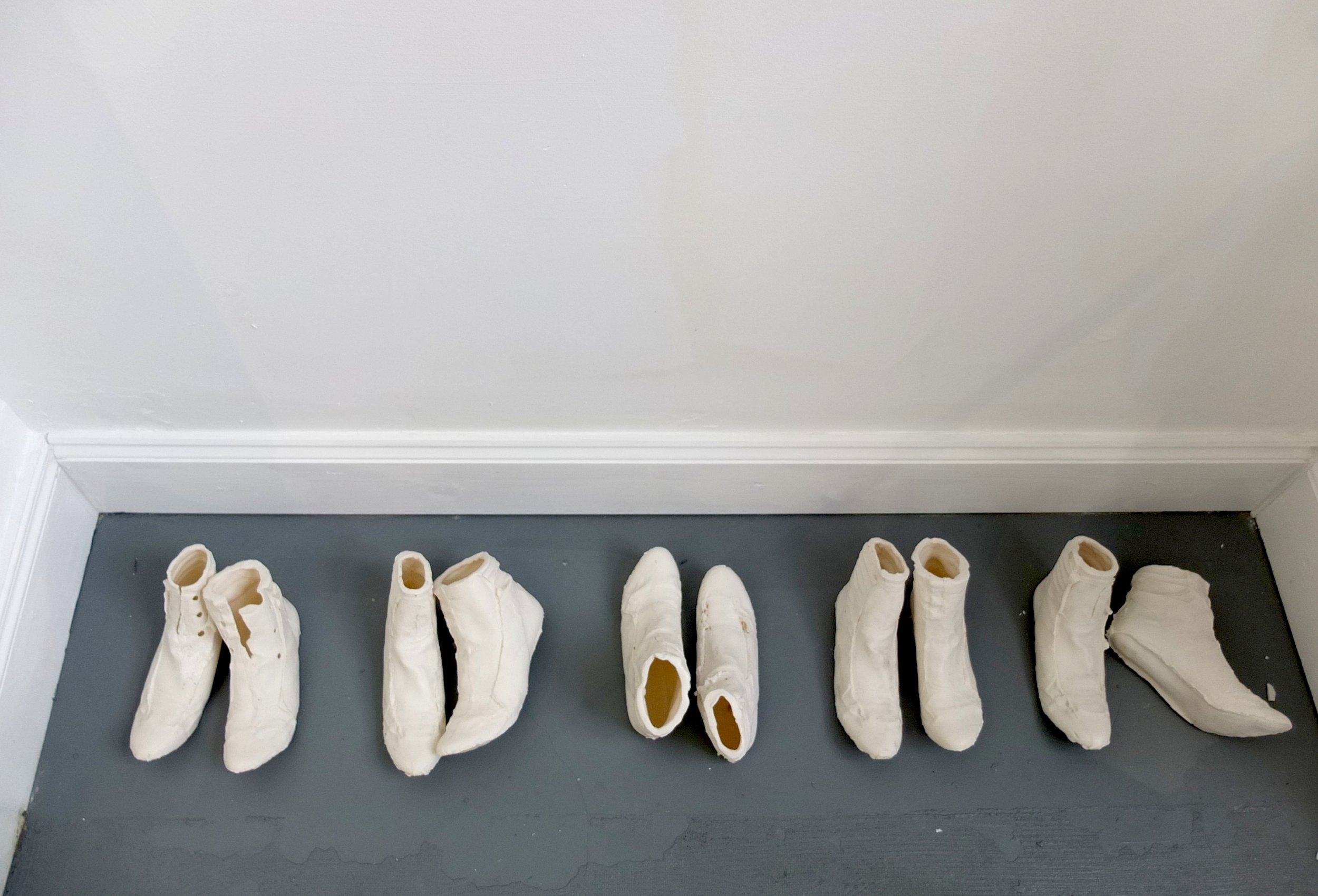 Rows of white plaster casts resembling shoes or boots on a dark floor against a plain white wall.