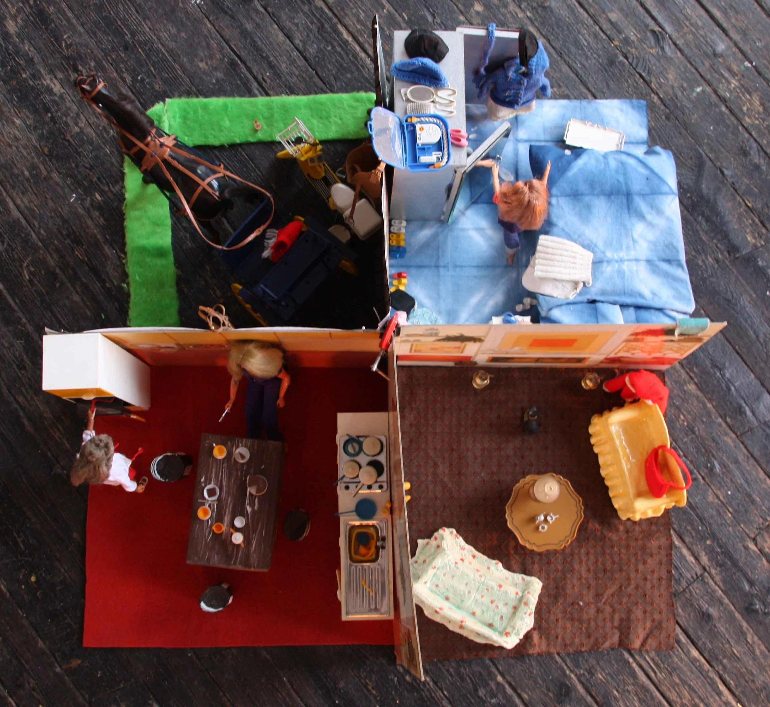 An overhead view of a miniature play kitchen setup with children playing, featuring a red table, toy kitchen appliances, and various toy food items on a dark wooden floor.