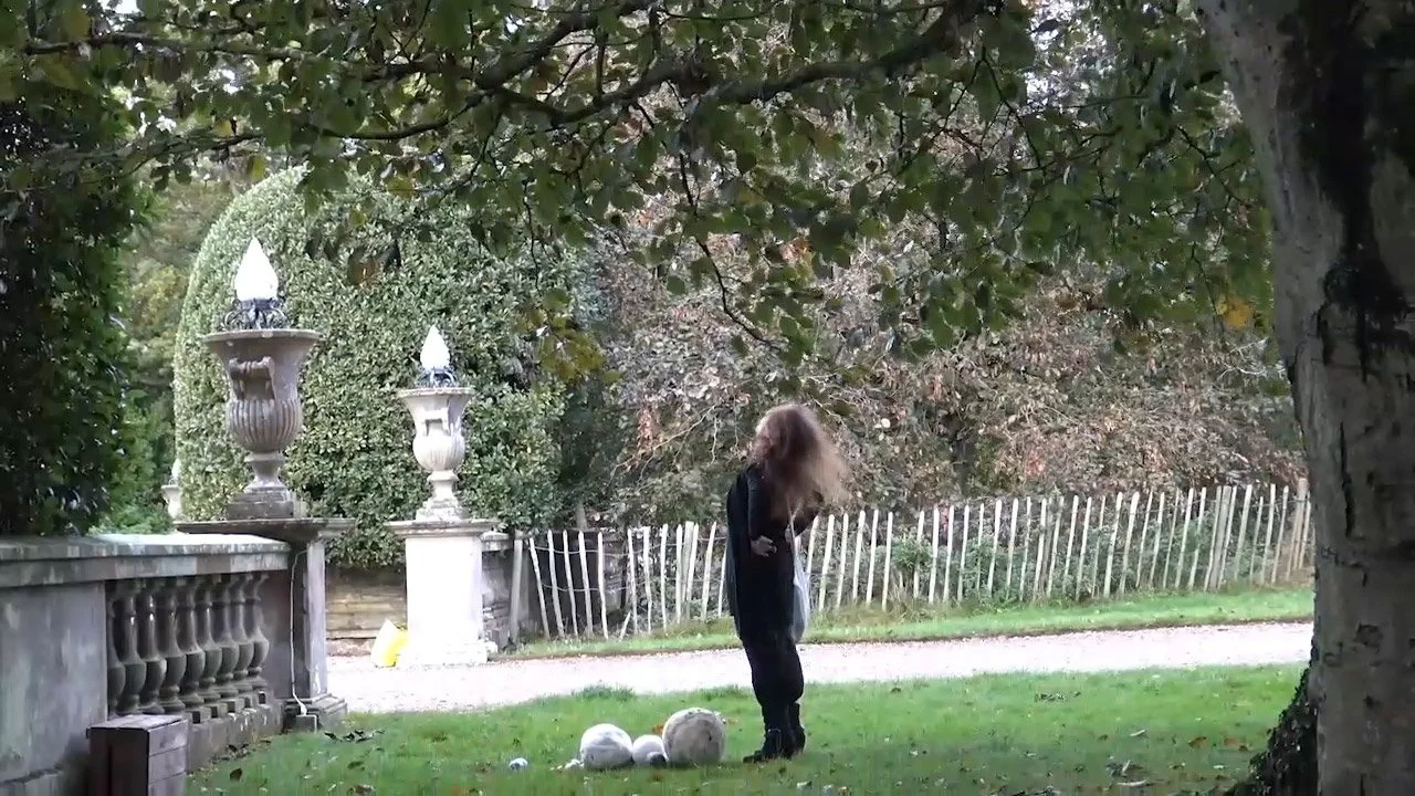 A woman with long hair stands on a grassy yard, stretching her back. There are three large decorative spheres on the ground nearby. A stone fence with ornate pillars and urns topped with lights is visible, alongside trees and plants.