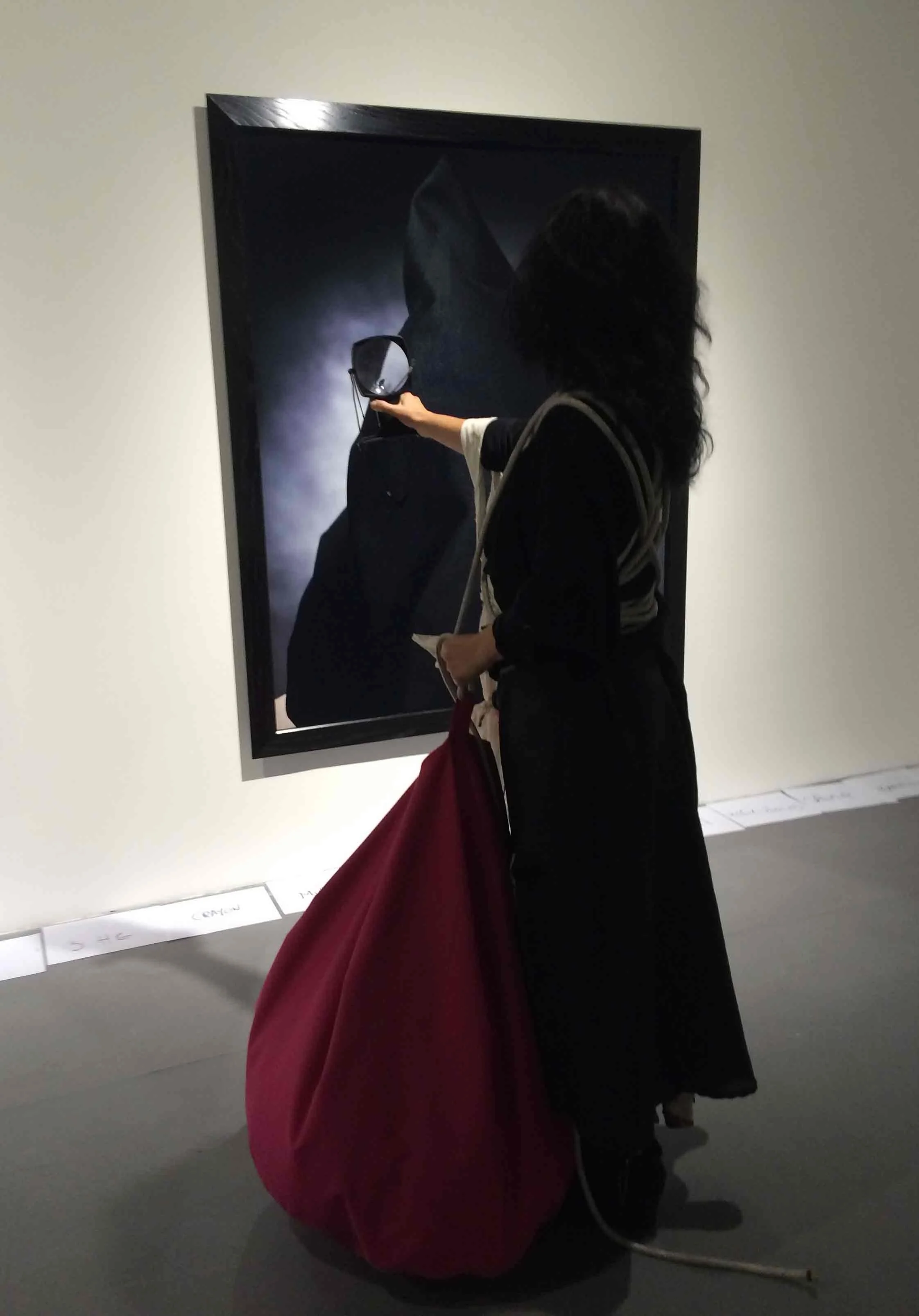 A woman in black attire with curly hair is holding a round mirror in front of a dark-hooded figure in a painting at an art gallery. A large crimson bag is hanging from her arm.