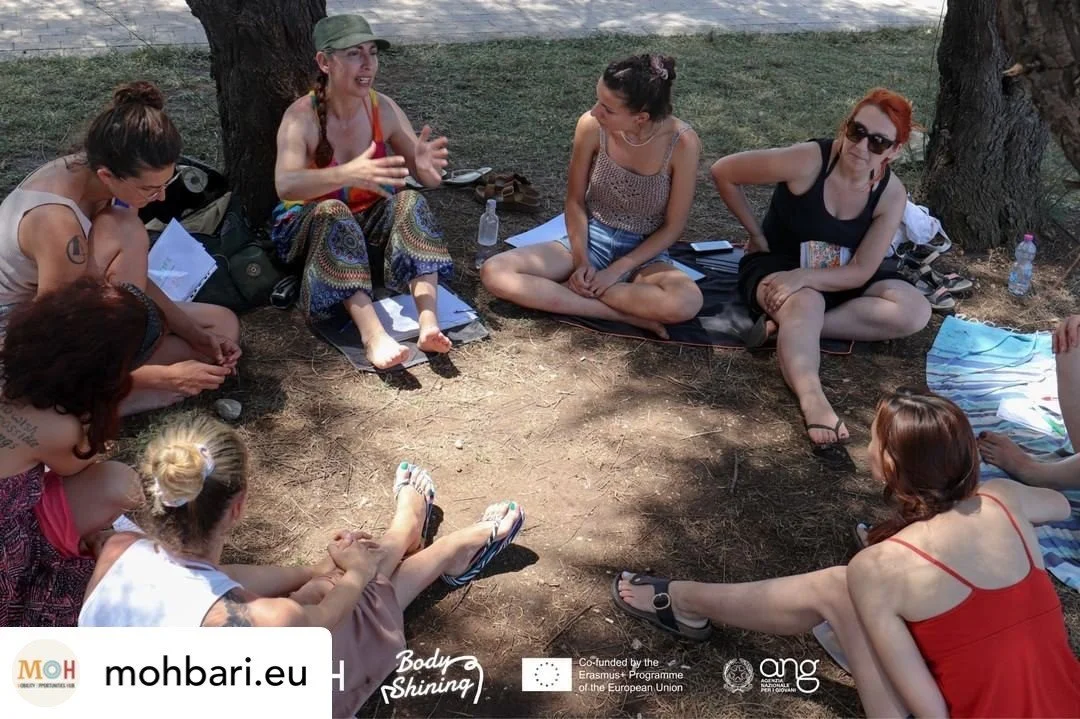 Group of women sitting cross-legged in a circle outdoors on a dirt ground, engaging in a discussion under a tree, with water bottles and notebooks nearby.