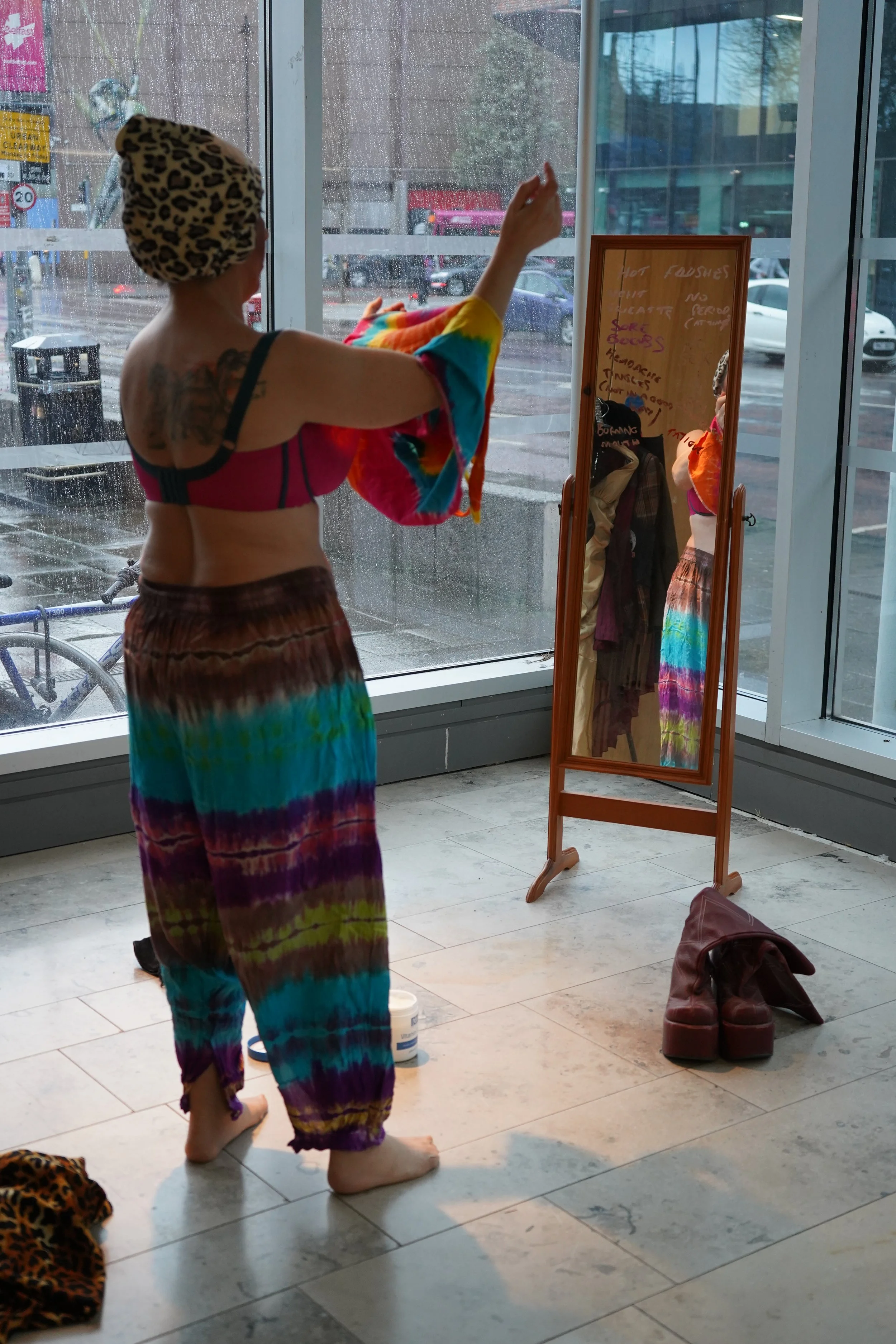 A woman standing indoors in front of a large window, wearing a colorful tie-dye skirt and a leopard print headwrap. She appears to be adjusting a rainbow-colored cloth or towel. There is a full-length mirror showing her reflection, a pair of brown bo