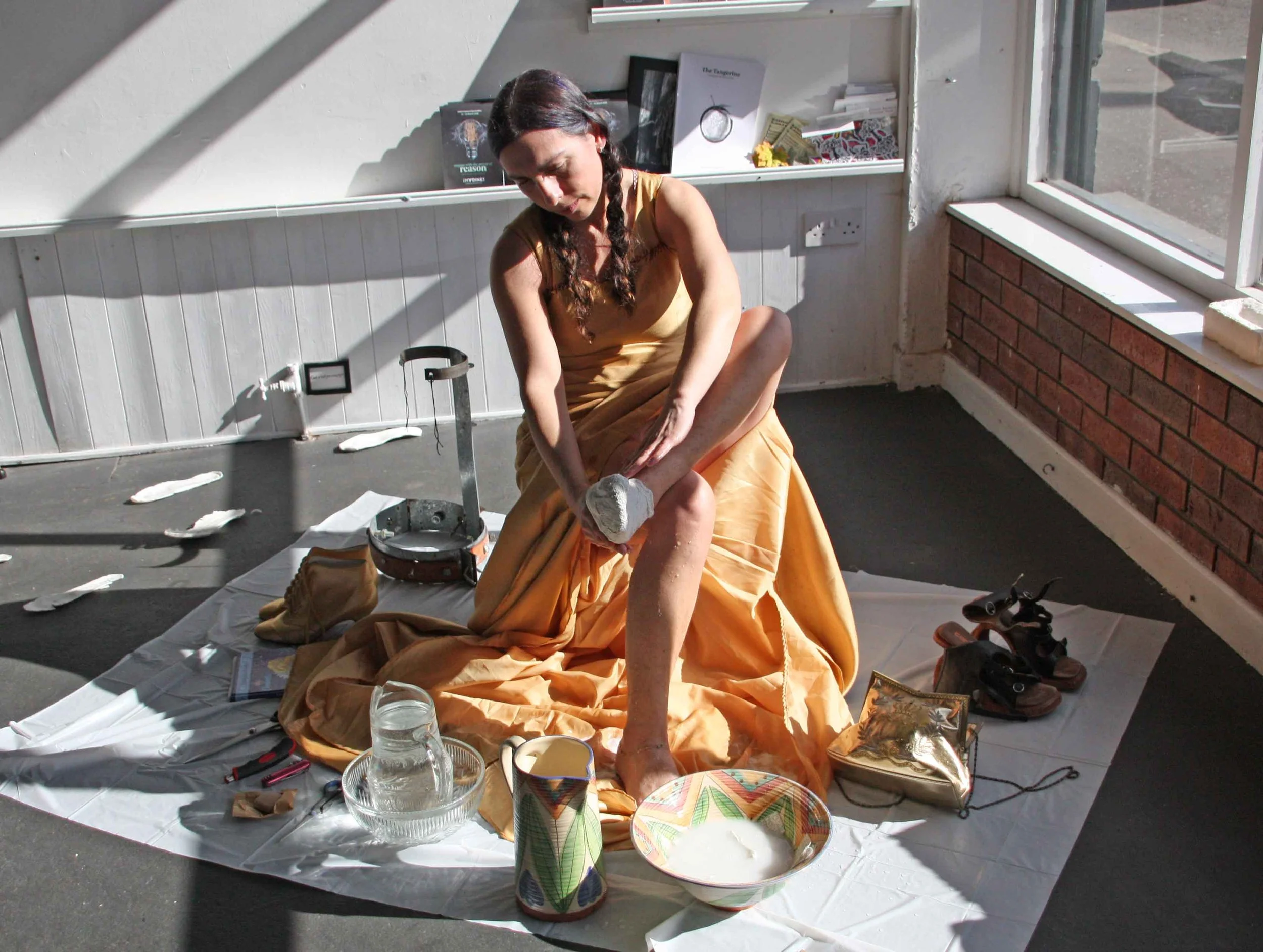 A woman dressed in a yellow sari, sitting on the floor, barefoot, with her legs crossed. She is bowing her head and appears to be performing a ritual or prayer. Various items such as a glass of water, a decorative bowl, candles, and other ceremonial 