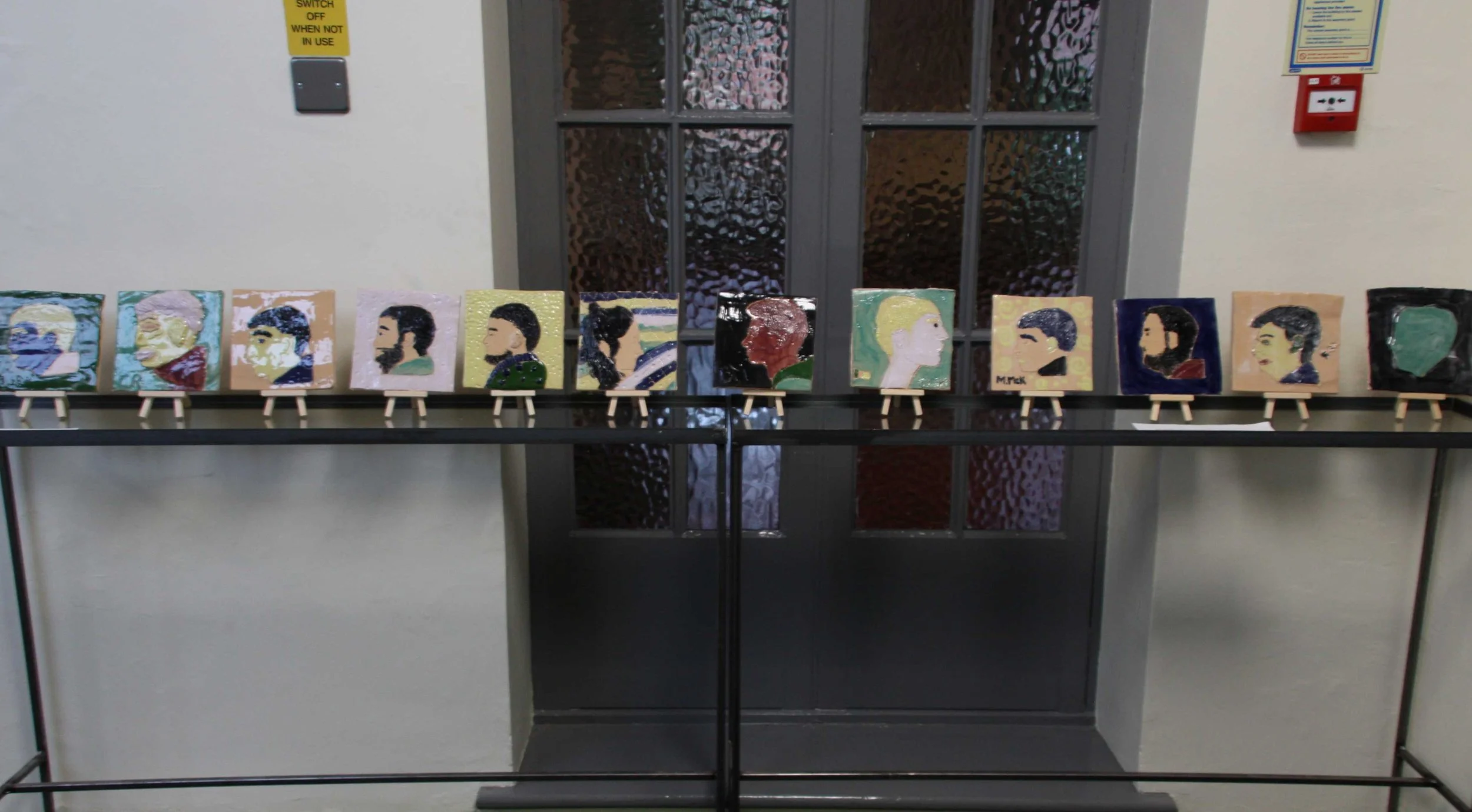 A row of ten painted portraits on small ceramic portrait tiles displayed on a table in front of a glass door with textured glass panes.