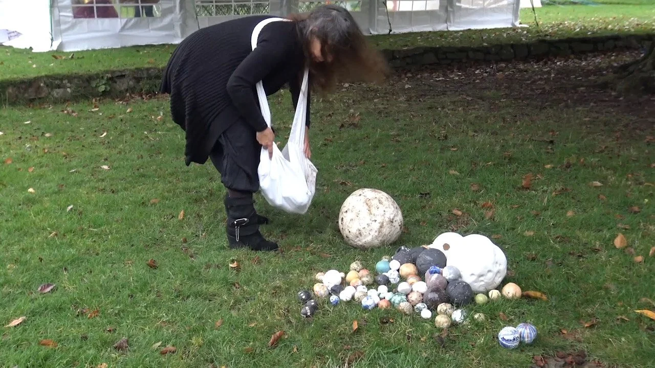 A woman with dark hair and black clothing crouches down on a grassy lawn, placing a white bag among a variety of decorative balls and sculptures on the ground. The background features a stone edge and a white fence.