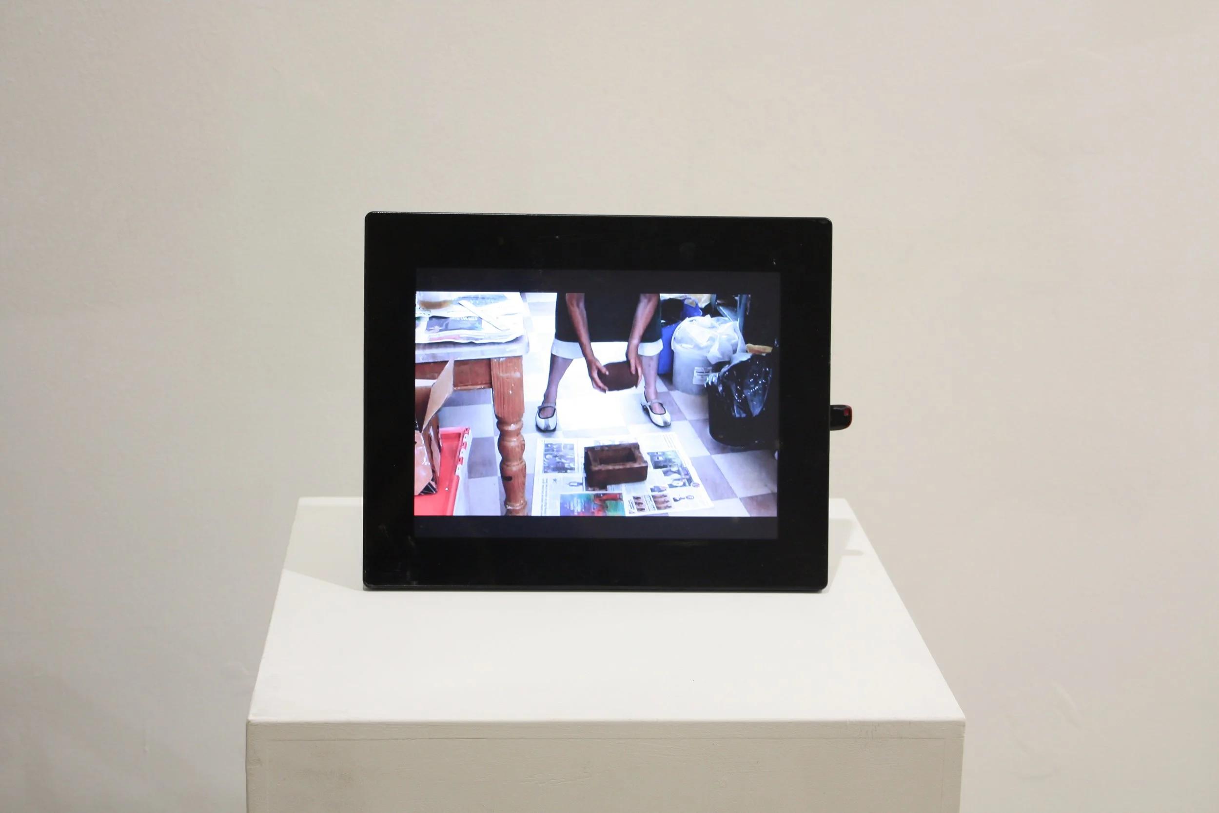 An art installation of a screen showing a person standing with their legs apart, holding an object between their legs, with a cluttered background of papers, bags, and furniture.