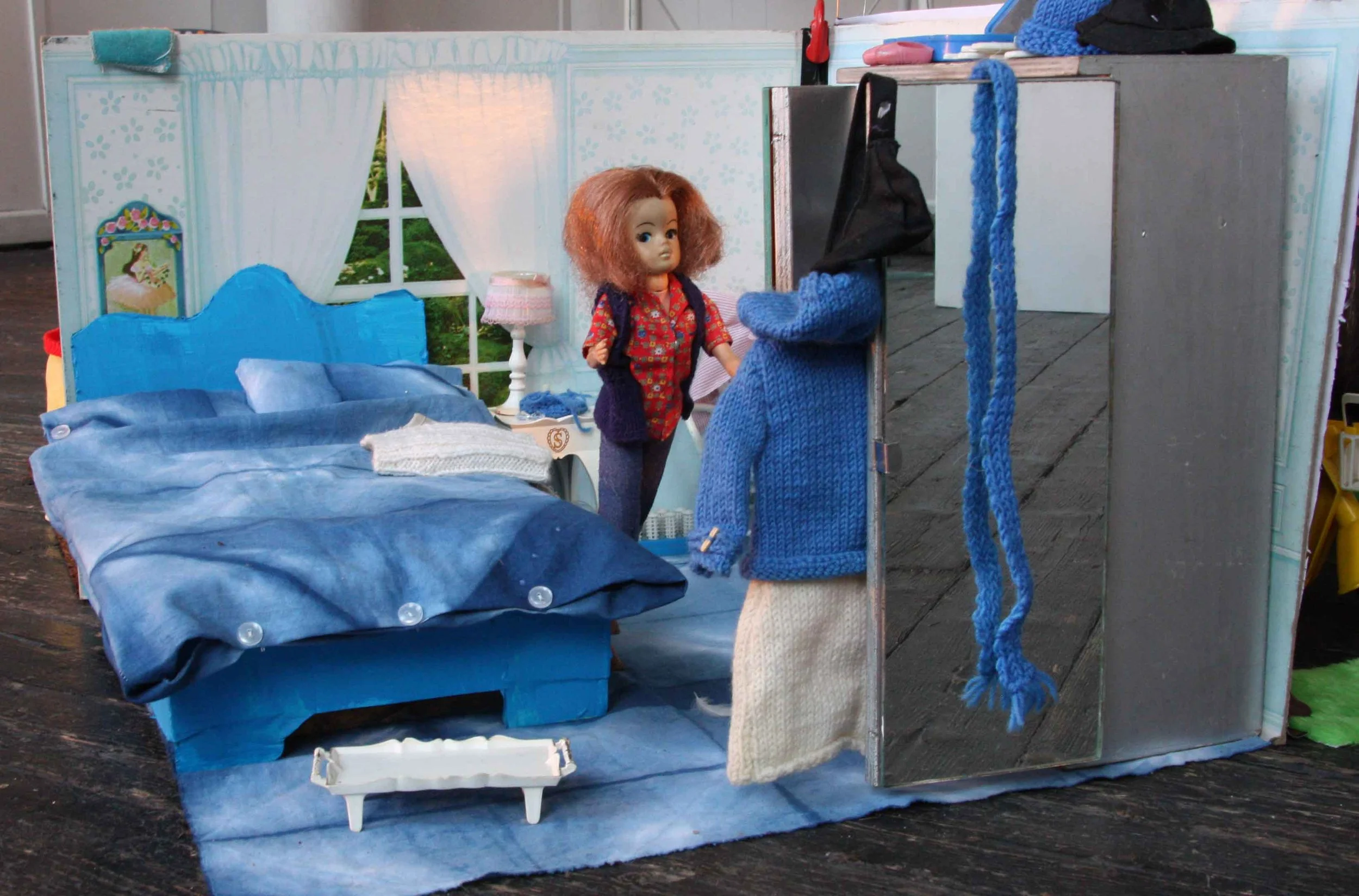 A miniature set resembling a cozy bedroom with a blue bed, window, and lamps, featuring two dolls with one wearing a colorful shirt and the other with a blue sweater, on a wooden floor.