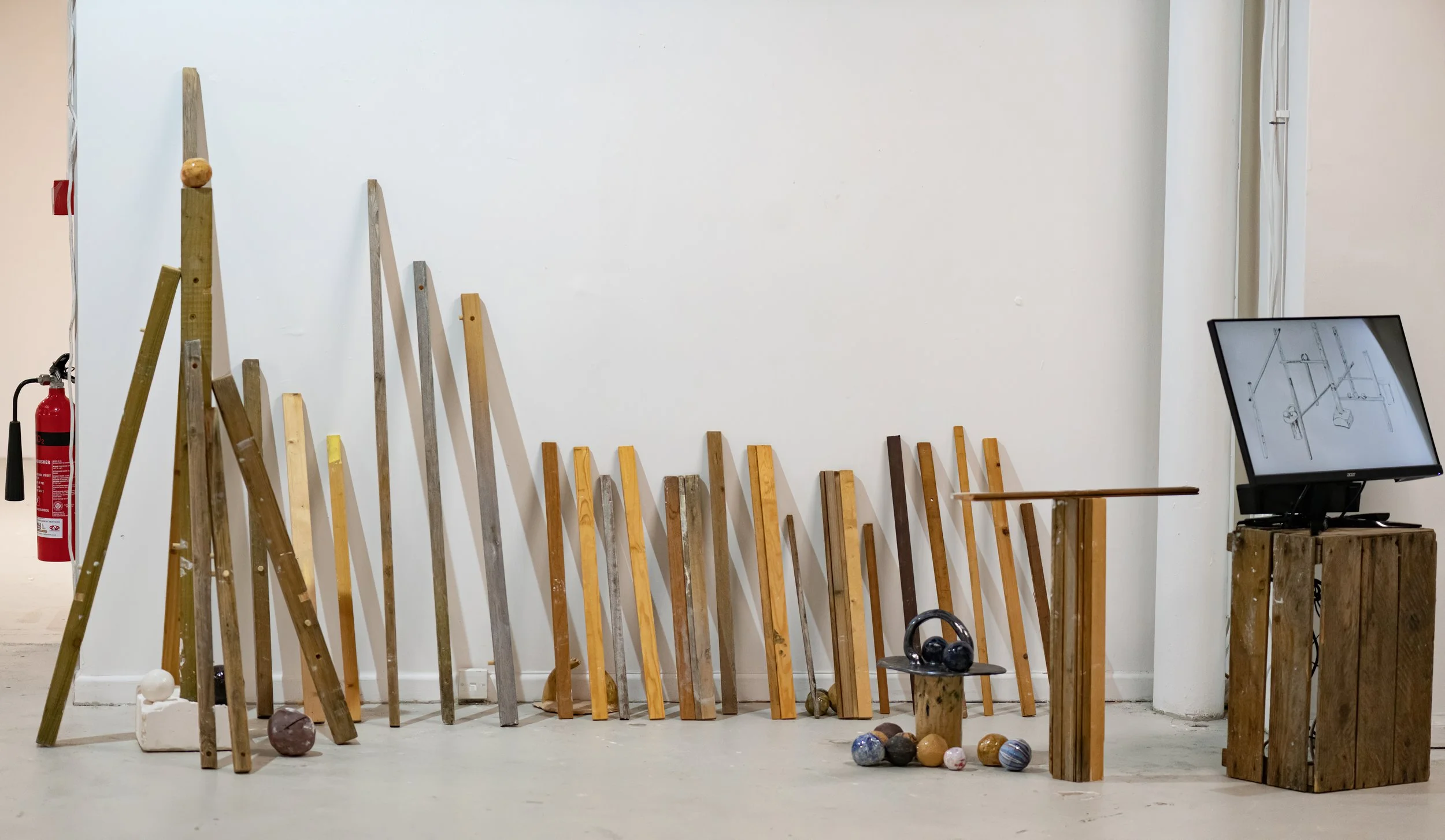 Rows of thin timber lined against a white wall in an indoor setting, with a television on a wooden stand to the right, playing technical drawings.
