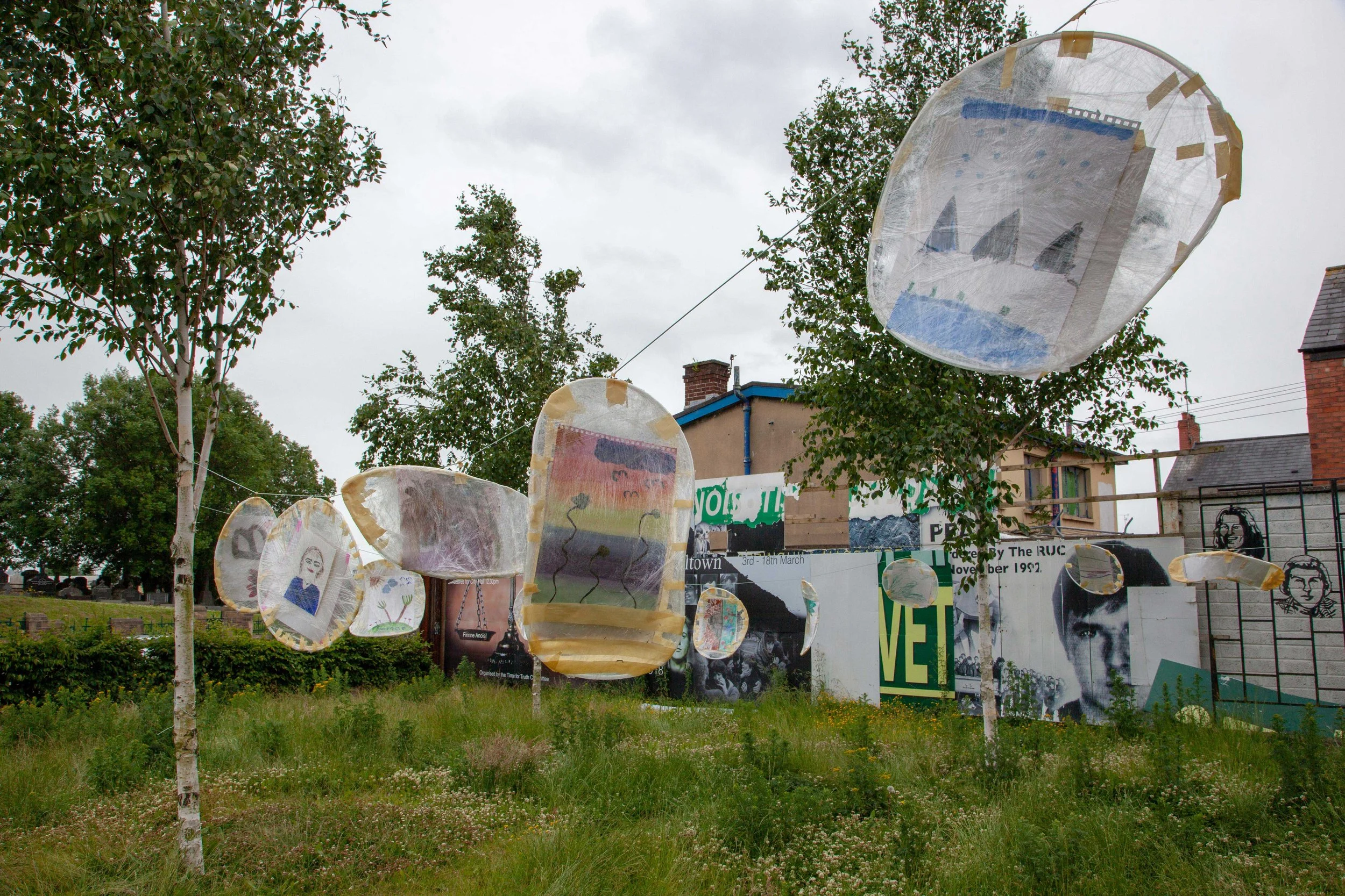 Outdoor art installation of several paper lanterns hanging on wires, with trees and a building in the background under a cloudy sky.