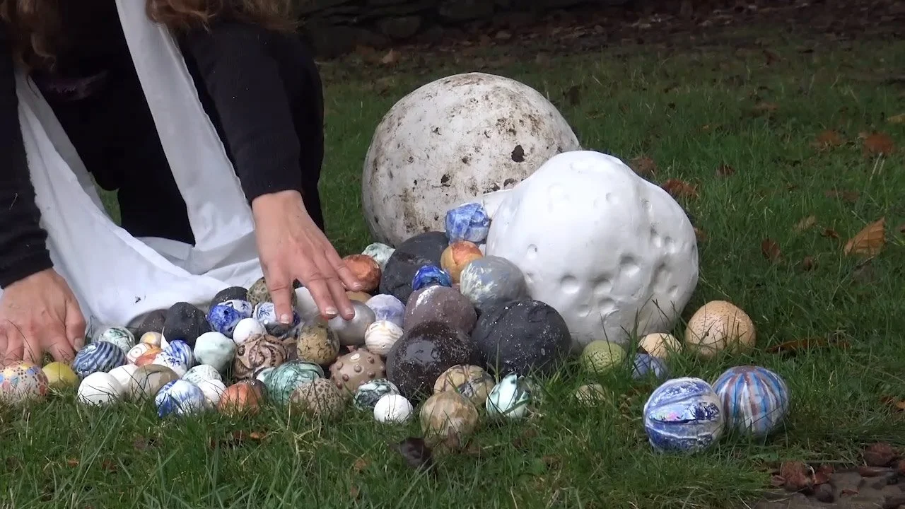 Person arranging decorative ceramic balls outdoors on grass with scattered leaves, variously colored and patterned balls, some large and textured.