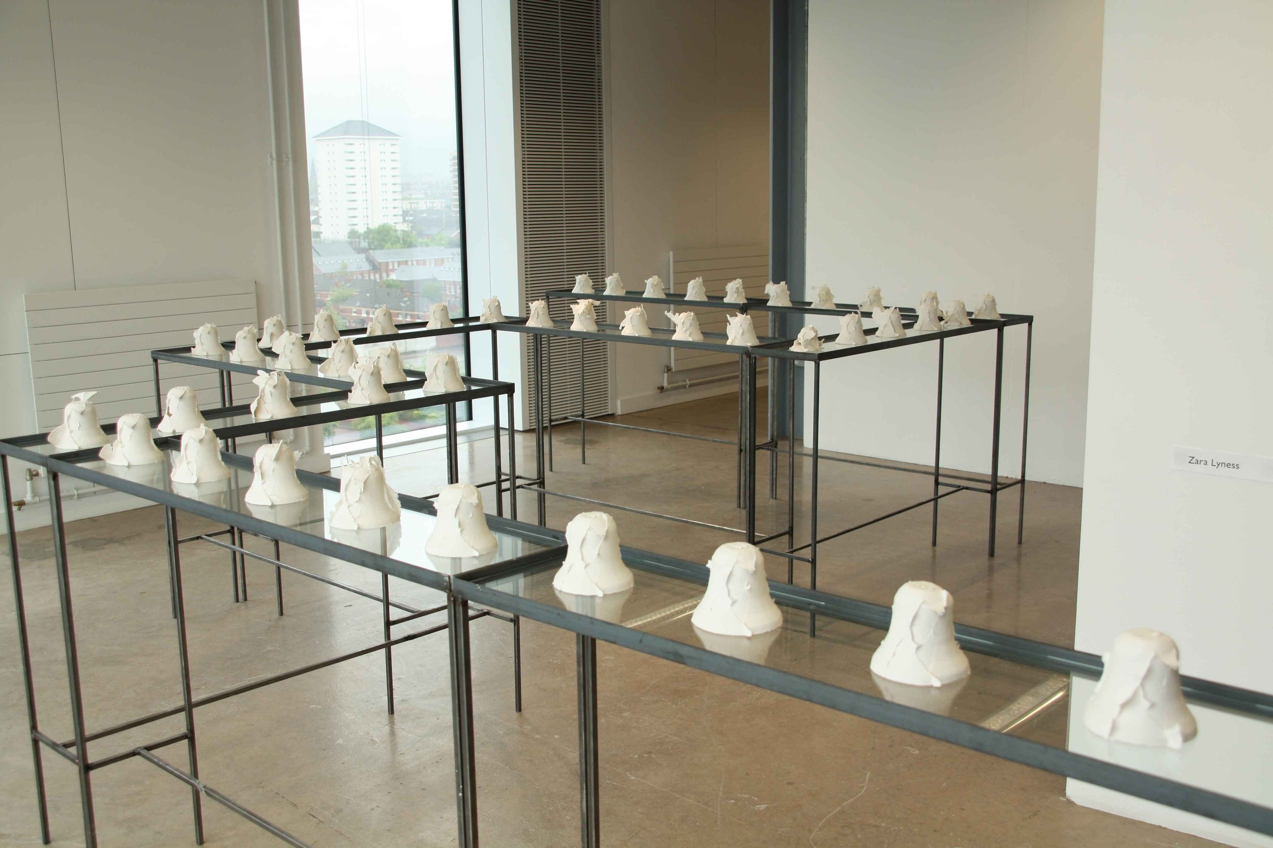 Contemporary art installation with white sculpted bells on black metal tables in a bright gallery space, with a large window overlooking a cityscape.