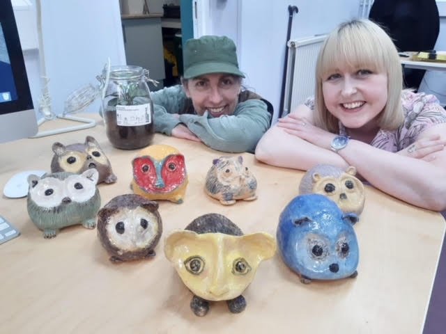 Two women are sitting at a table with nine small hedgehog ceramic or clay sculptures, which include owls, a bear, a fox, and other animals, along with a jar filled with dark substance, in a room with white walls and a radiator.