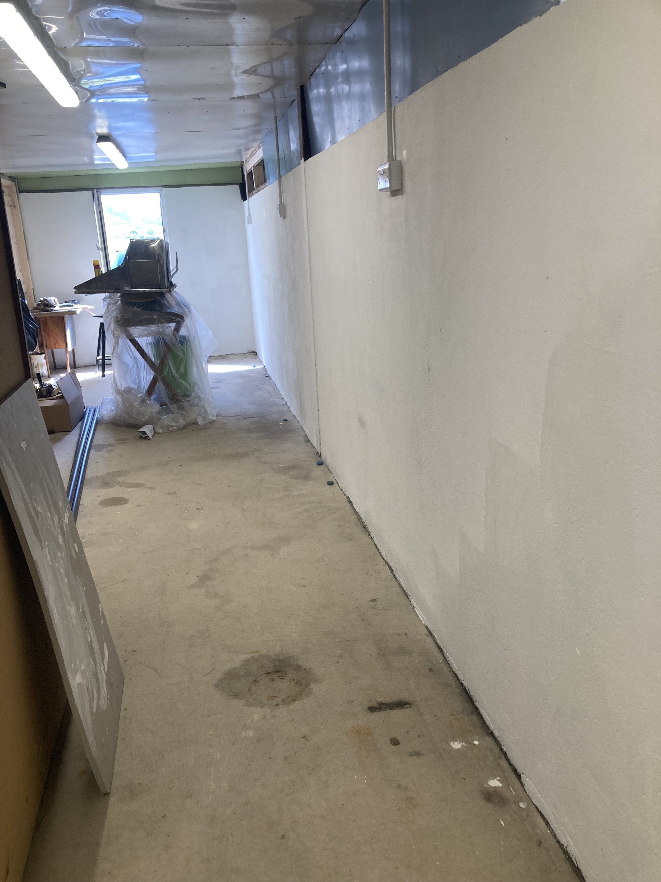 A hallway under construction with a white wall on the right, a yellow door on the left, and construction materials scattered on the floor. There is a window at the end of the hallway letting in natural light.