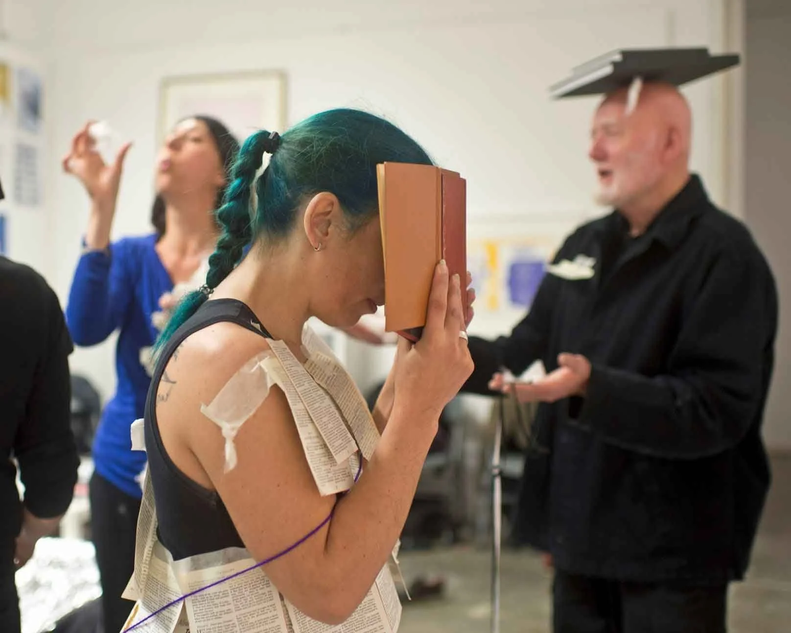 A woman with teal-colored hair and tattoos on her arms is holding a closed book in front of her face. She has taped pages to her arms and torso and is wearing a sleeveless top. In the background, a woman in a blue jacket and a man in a black uniform with a graduation cap and diploma are visible, suggesting a stressful or emotional event, possibly a graduation or ceremony.