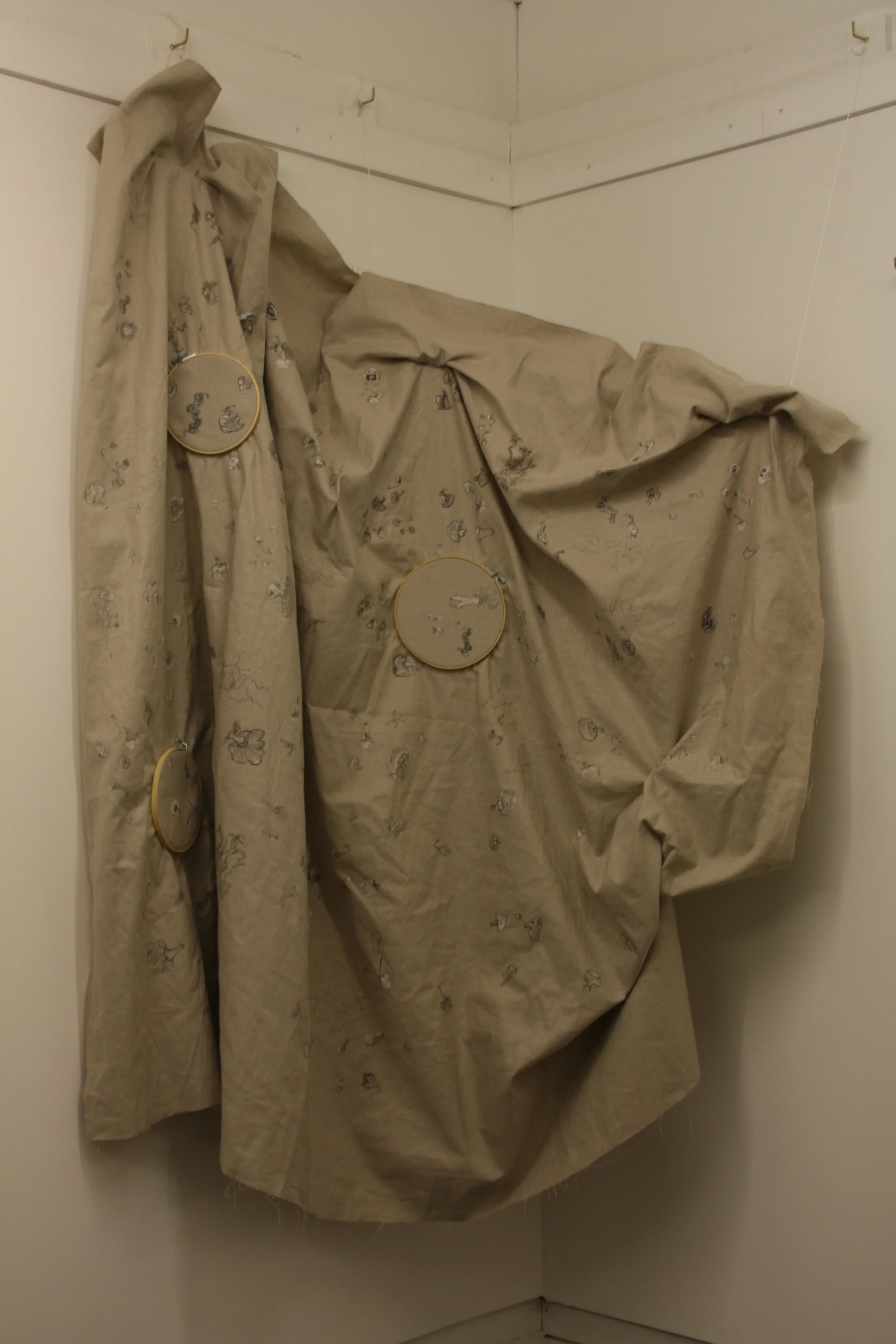 A corner of a room with beige fabric draped over rods, with embroidery of small animals, possibly rabbits or mice, embroidered on the fabric.
