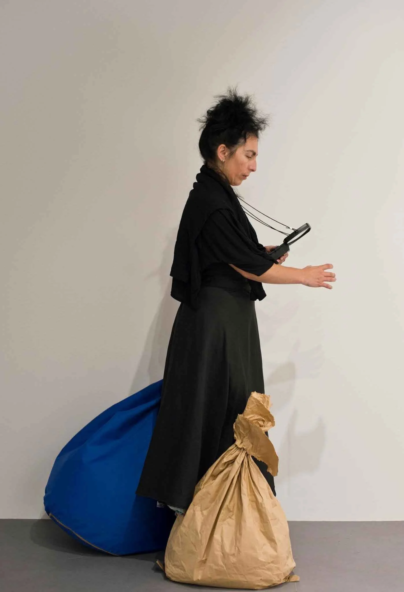 A woman with spiky black hair wearing black clothing is standing against a plain white wall, looking at her phone. She has a blue bag on the floor next to her and a brown paper bag at her feet.