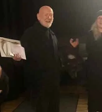 Older man with a beard, wearing a black shirt, smiling and standing indoors.