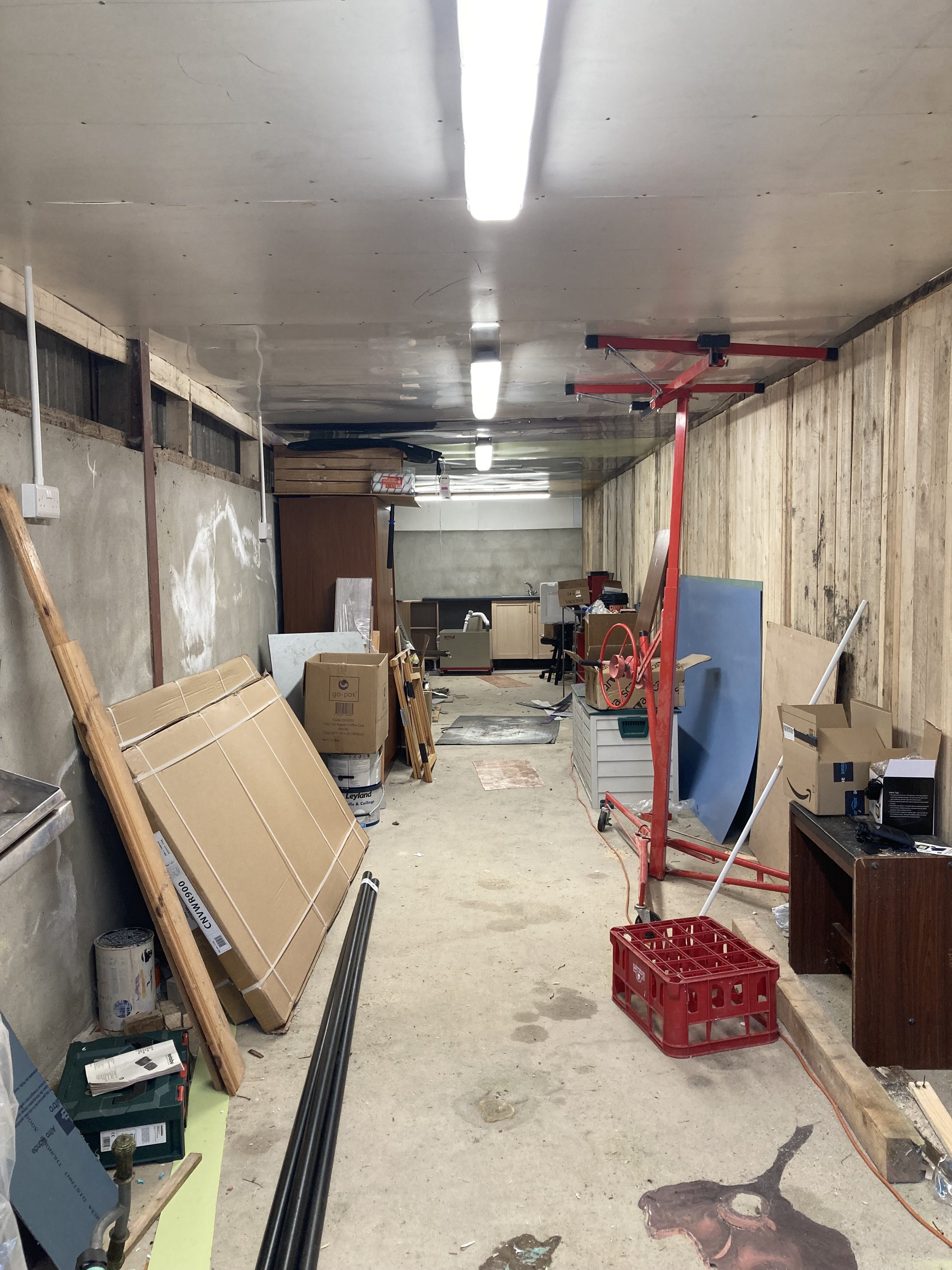 A cluttered workshop or storage room with various tools, boxes, and materials, including a red scaffold, wooden panels, and cardboard boxes, with fluorescent lights on the ceiling and a concrete floor.