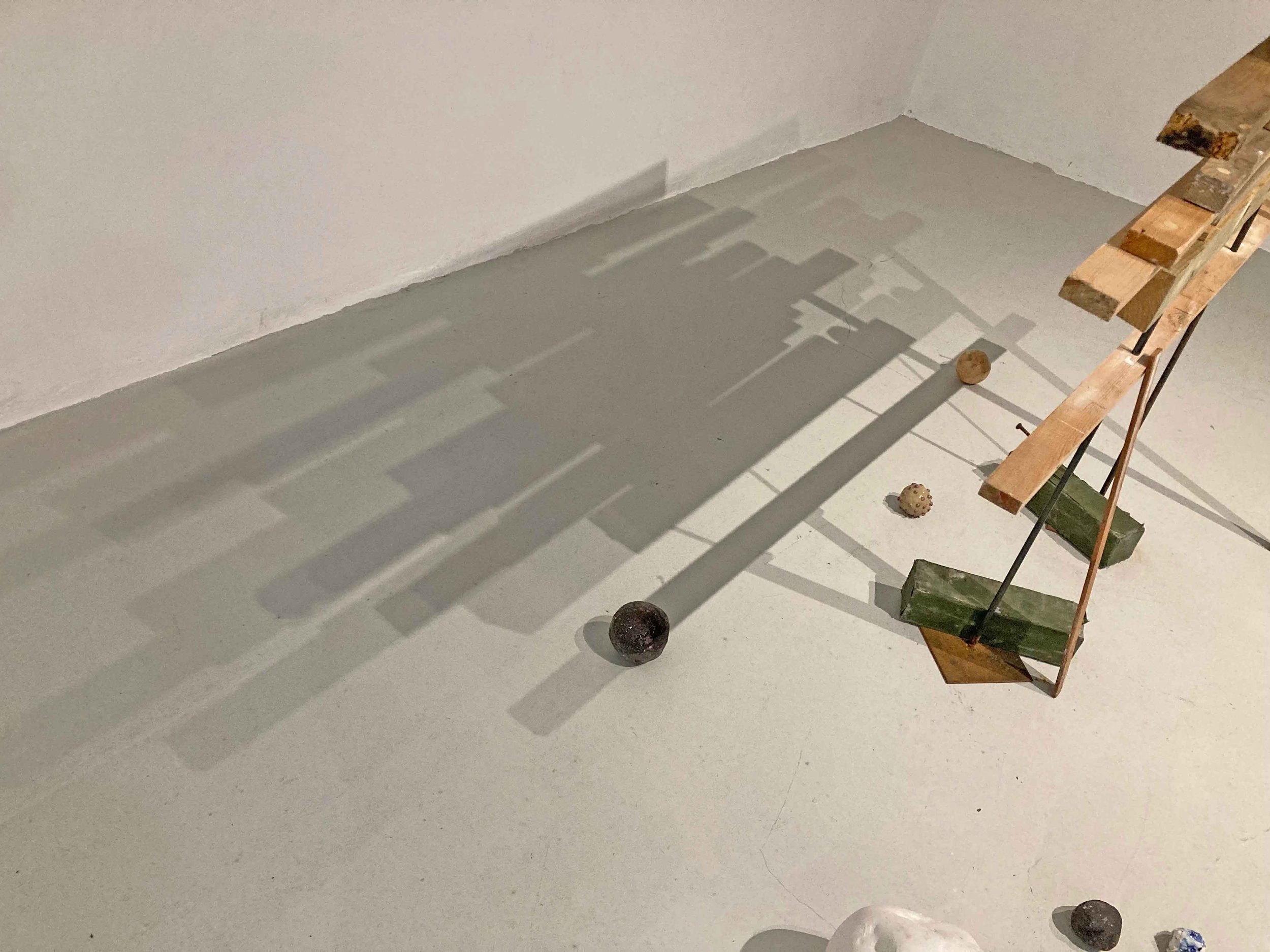 Construction in a gallery of  a partially assembled wooden structure and three spherical objects on the floor, casting shadows on the wall.