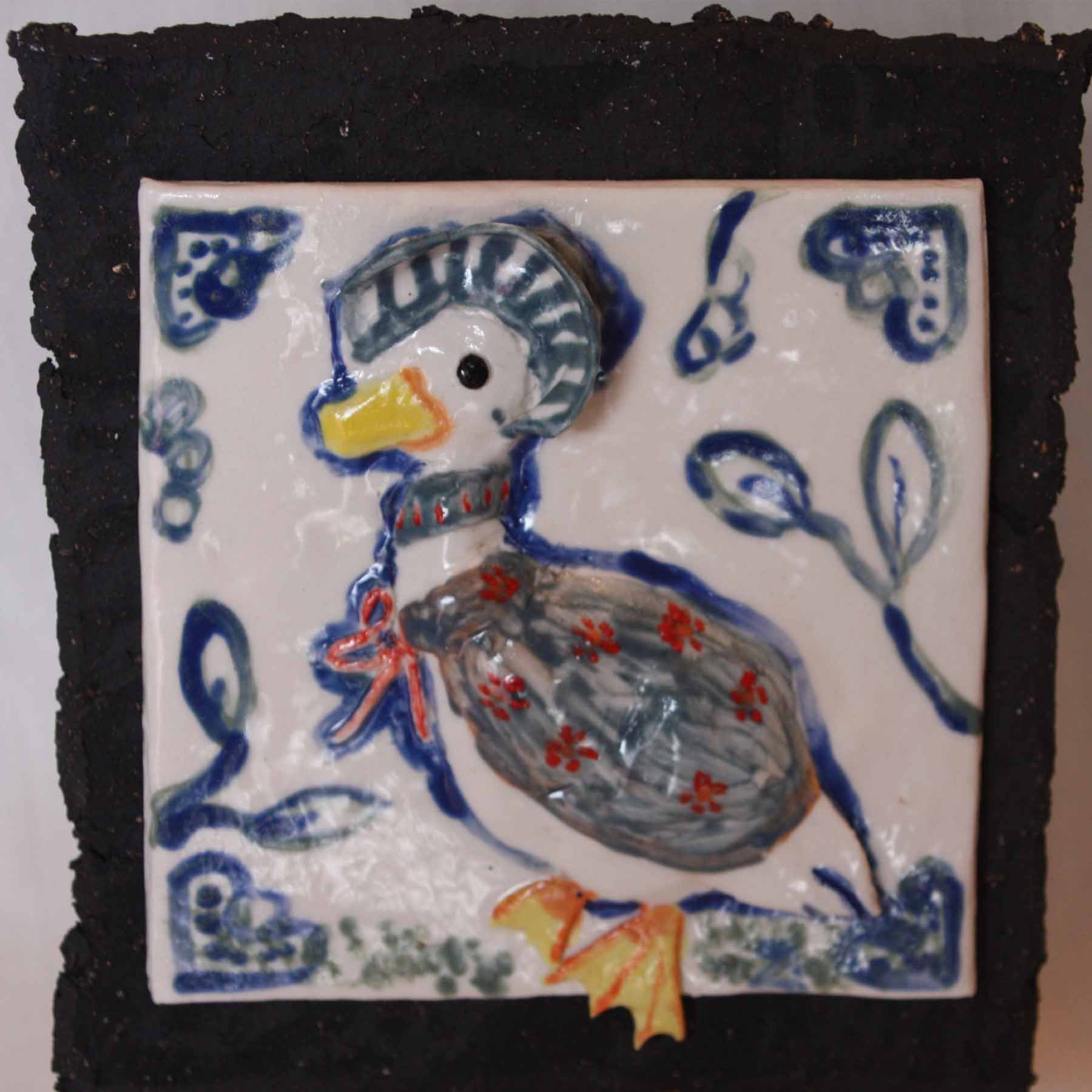 Ceramic tile artwork featuring a duck with a gray body, yellow beak, and black eye, surrounded by blue decorative patterns and a rural scene with a duck standing on grass.
