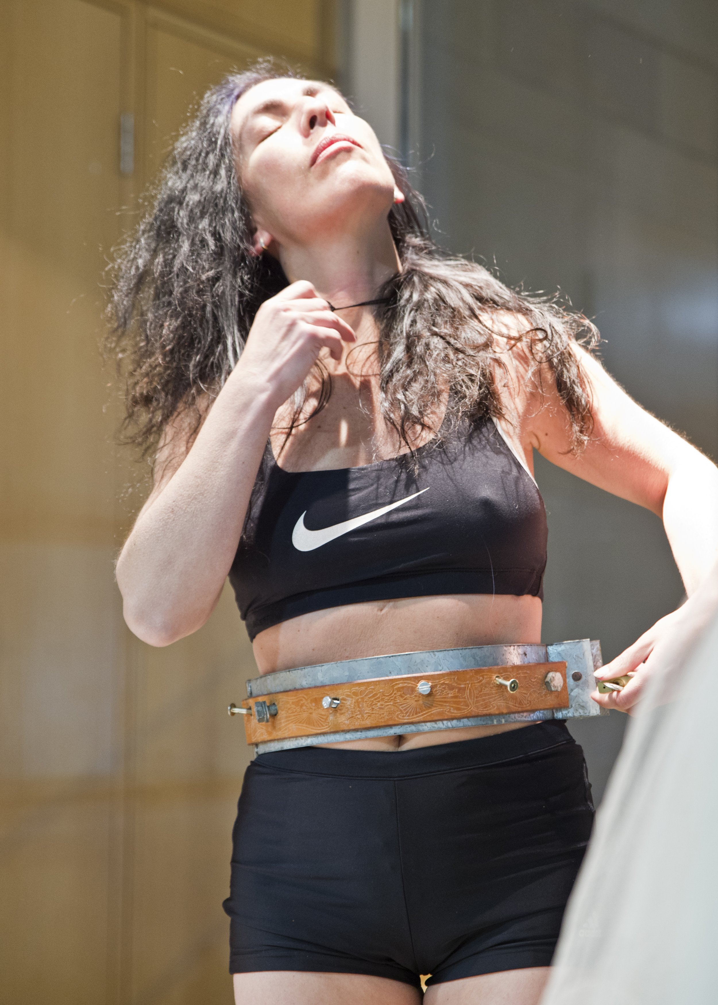 A woman with long, curly dark hair in athletic clothing, including a black Nike sports bra, appears to be in a sports lab or recovery room, with her eyes closed and head tilted back. She is holding a thin necklace in her right hand and is wearing a m