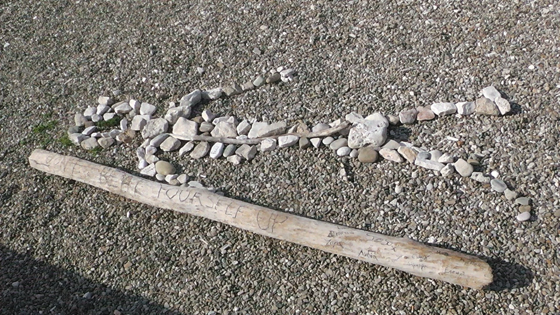 A pebble-covered beach with a large white wooden stick inscribed with the words names and small rocks arranged in a semi-circle.